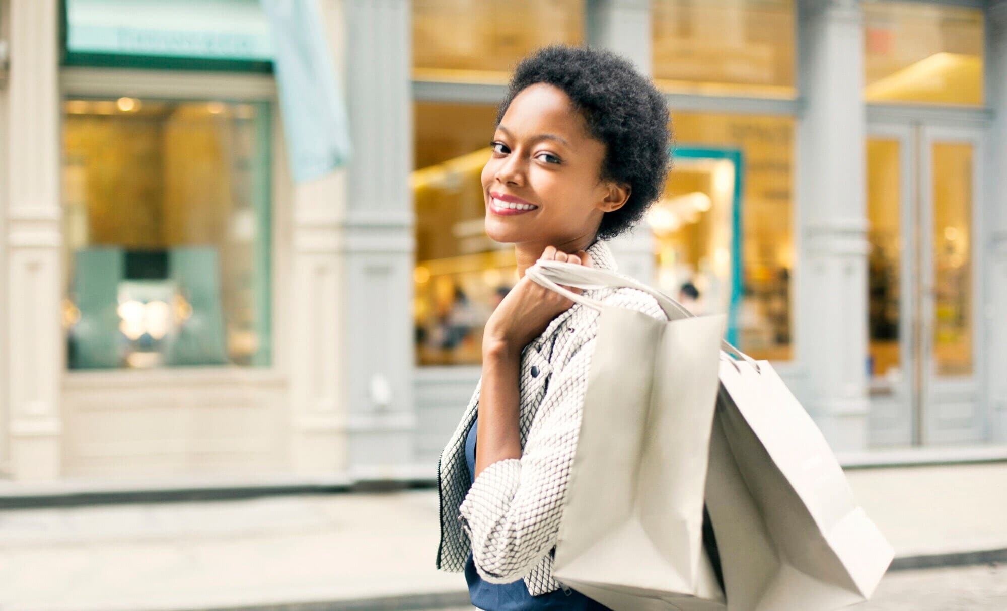 Eine junge Frau vor einem luxuriösen Geschäft trägt zwei Einkaufstüten über der Schulter und lächelt glücklich in die Kamera Eine junge Frau vor einem luxuriösen Geschäft trägt zwei Einkaufstüten über der Schulter und lächelt glücklich in die Kamera