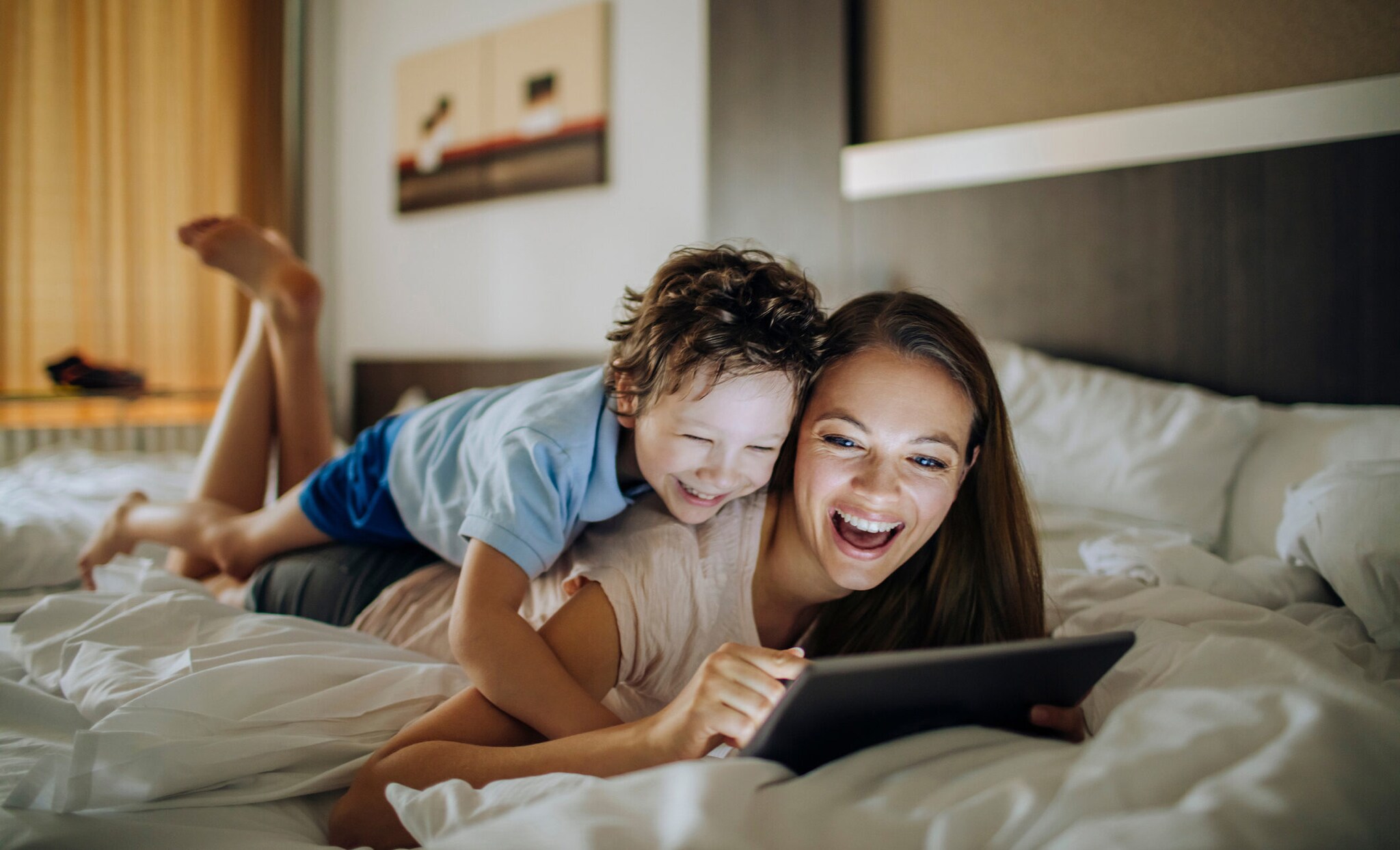 Eine Mutter und ihr kleiner Sohn liegen aneinander gekuschelt im Bett und schauen lachend auf ein Tablet