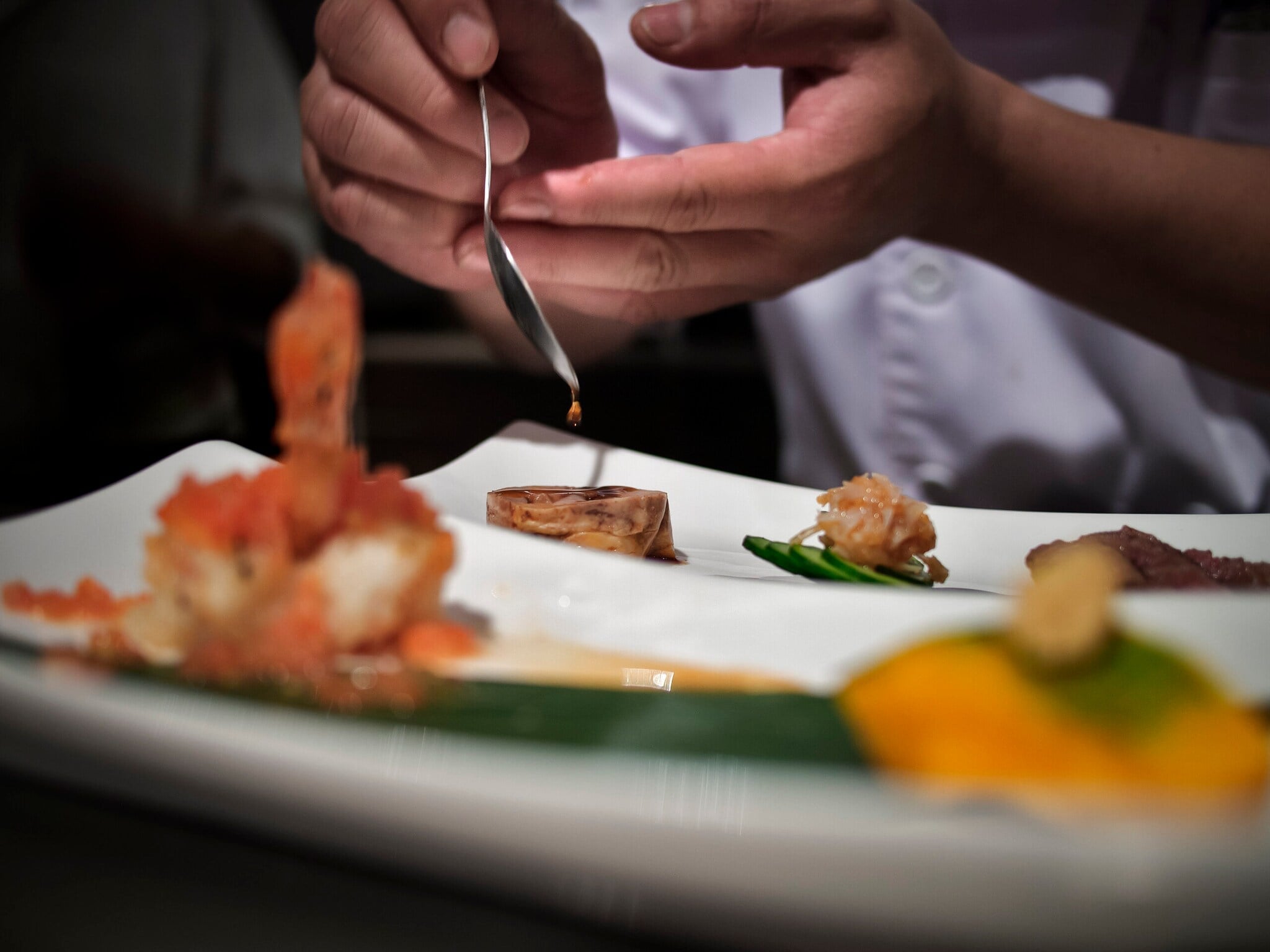 Die Hand eines Küchenchef beträufelt mit einem Löffel ein exquisites Gericht auf einem weißen Teller Die Hand eines Küchenchef beträufelt mit einem Löffel ein exquisites Gericht auf einem weißen Teller