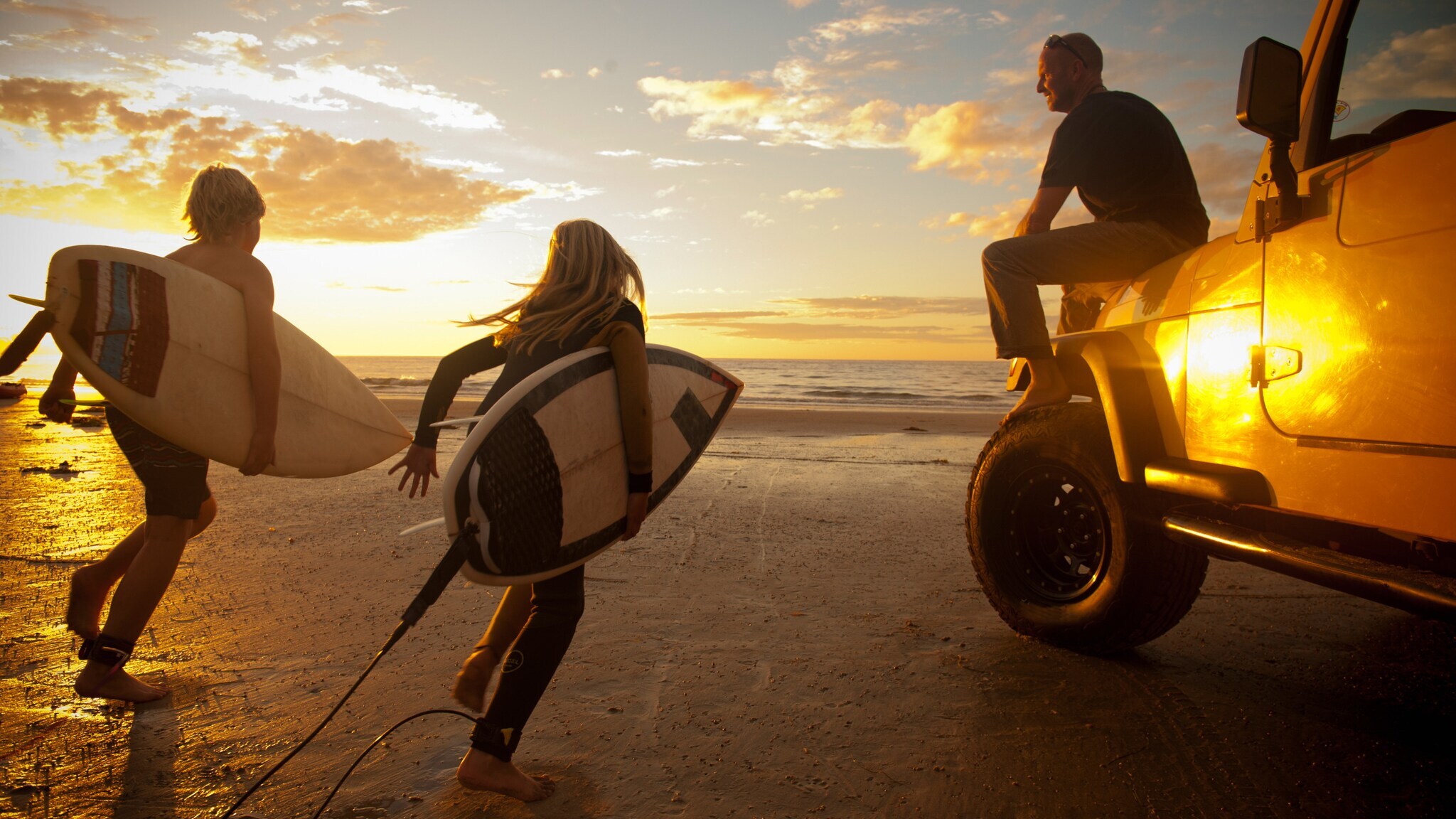 Rückansicht von zwei Kindern mit Surfbrettern neben einem sitzenden Mann auf der Motorhaube eines Jeeps am Strand bei Sonnenuntergang.