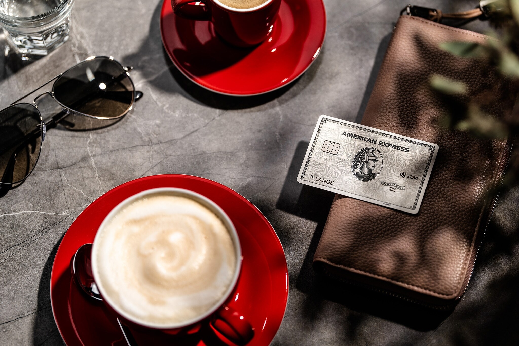 Eine Amex Platinum Card liegt auf einer Geldbörse auf einem Tisch neben einer roten Tasse mit Milchkaffee im Sonnenschein