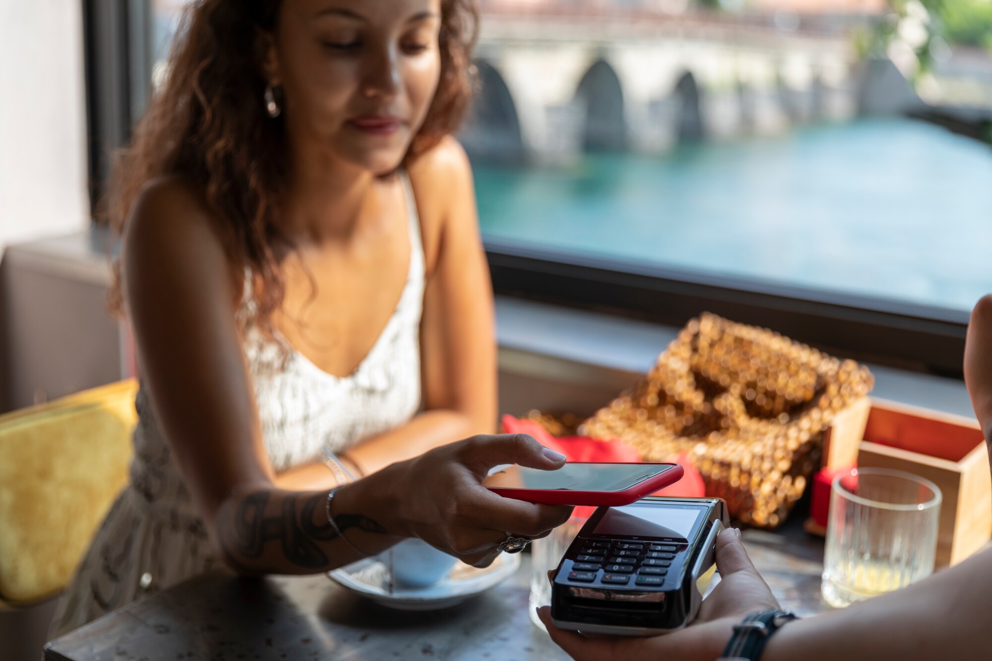 Eine junge Frau im Sommerkleid bezahlt kontaktlos mit ihrem Smartphone in einem Café an einem Fluss