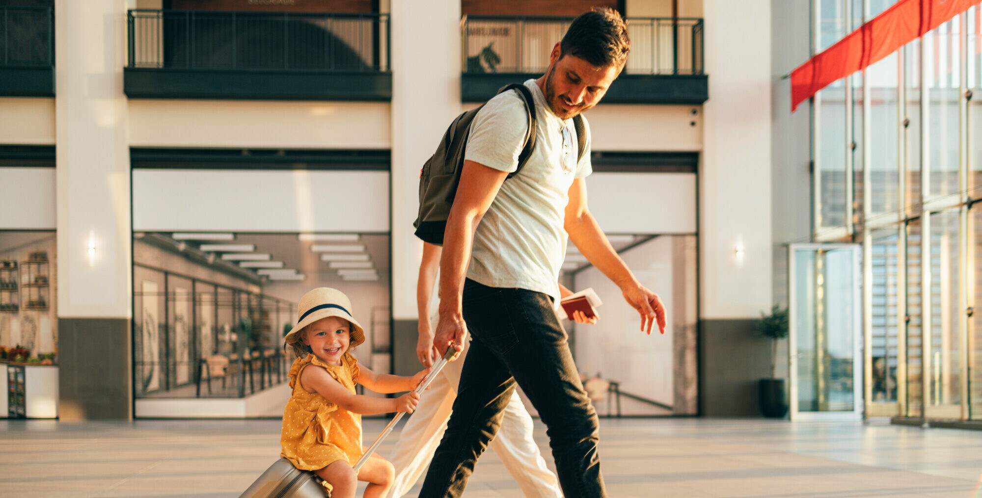 Ein kleines Mädchen im gelben Sommerkleid sitzt lachend auf einem Koffer, der von einem Mann durch eine Flughafenhalle gezogen wird Ein kleines Mädchen im gelben Sommerkleid sitzt lachend auf einem Koffer, der von einem Mann durch eine Flughafenhalle gezogen wird