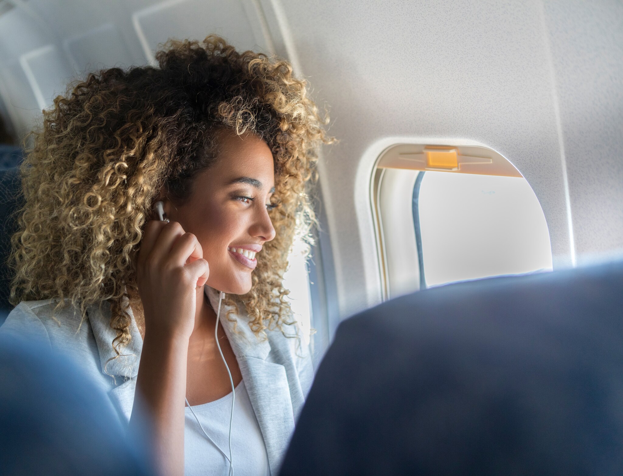 Nahaufnahme einer jungen, lächelnden Frau mit Kopfhörern auf einem Fensterplatz in einem Flugzeug während der Reise