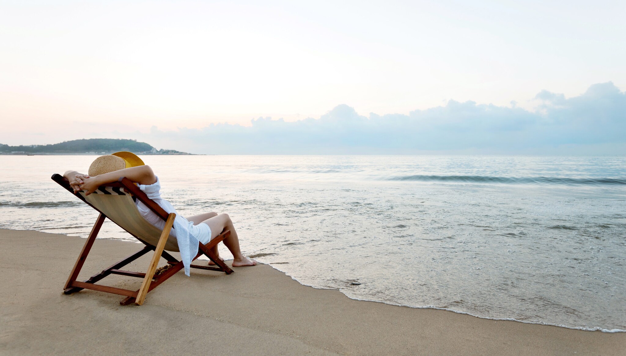 Rückansicht einer Frau mit Sonnenhut in einem Liegestuhl am Wasser an einem Sandstrand