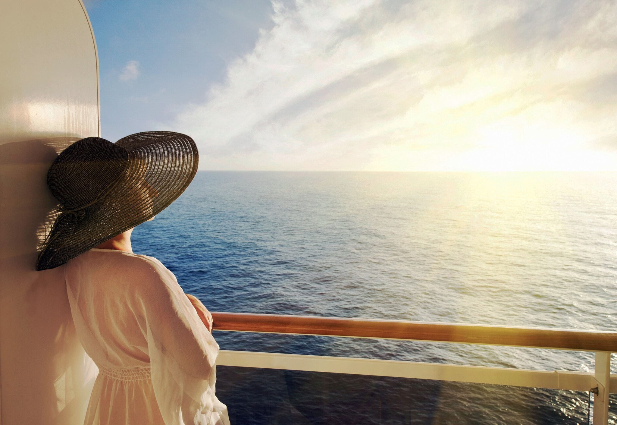 Eine elegante Frau mit Sonnenhut schaut im Sonnenuntergang von einem privaten Balkon eines Kreuzfahrtschiffes auf das offene Meer hinaus Eine elegante Frau mit Sonnenhut schaut im Sonnenuntergang von einem privaten Balkon eines Kreuzfahrtschiffes auf das offene Meer hinaus