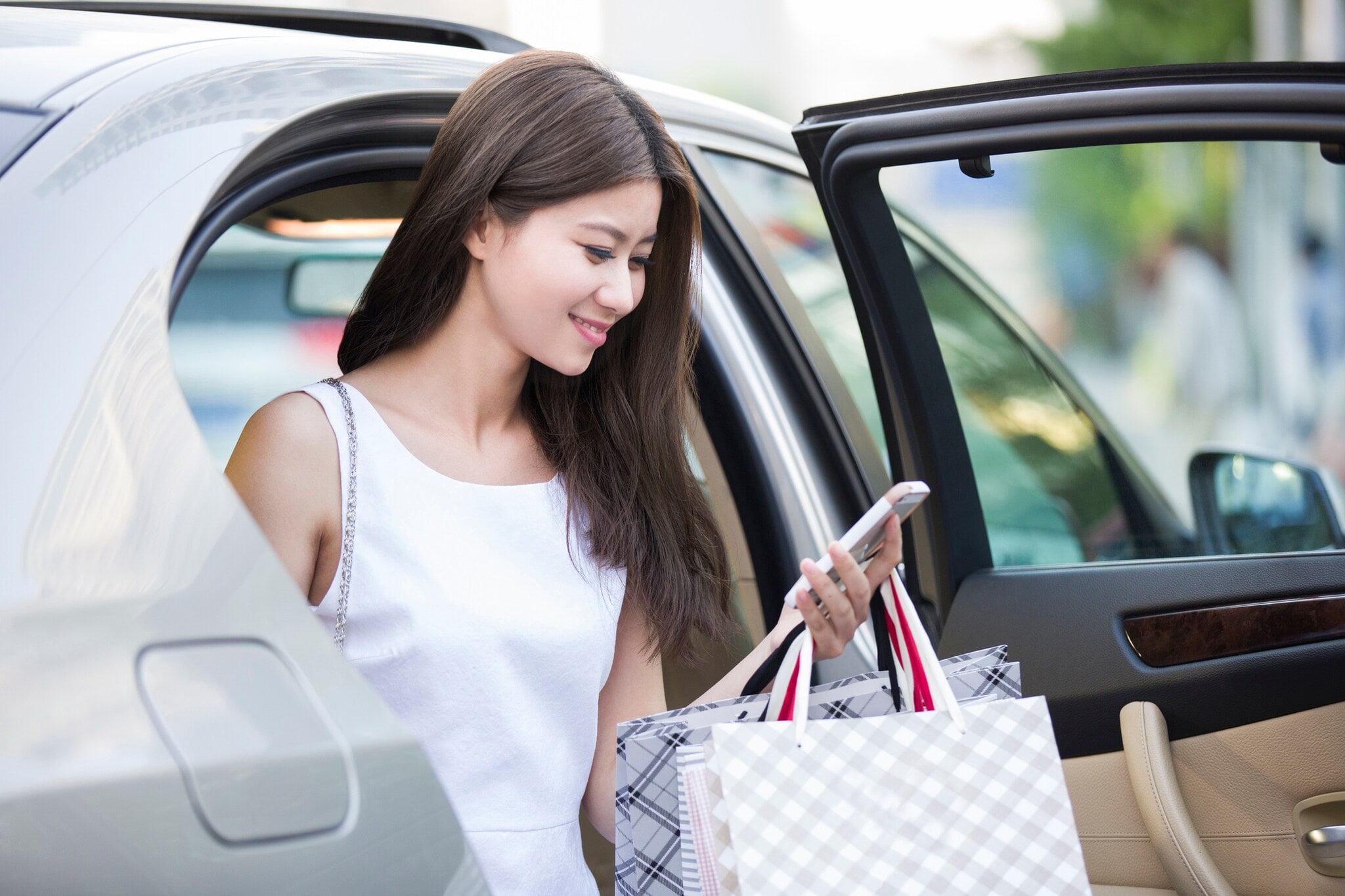 Eine elegante, junge Frau mit Einkaufstaschen in der Hand steigt aus einem Auto und schaut lächelnd auf ihr Smartphone Eine elegante, junge Frau mit Einkaufstaschen in der Hand steigt aus einem Auto und schaut lächelnd auf ihr Smartphone