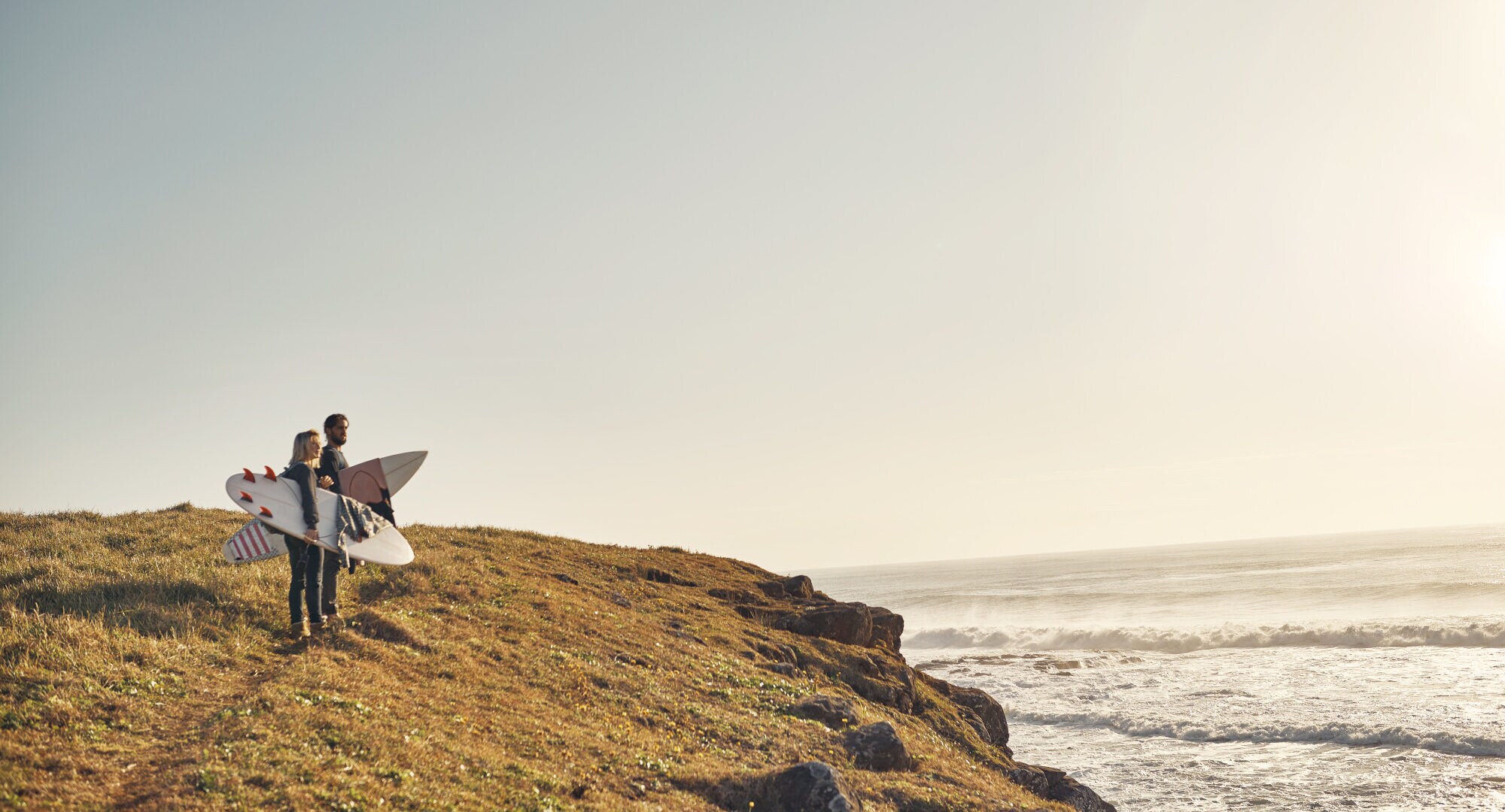 Zwei Personen mit Surfbrettern in der Hand schauen an einer Klippe aufs Meer