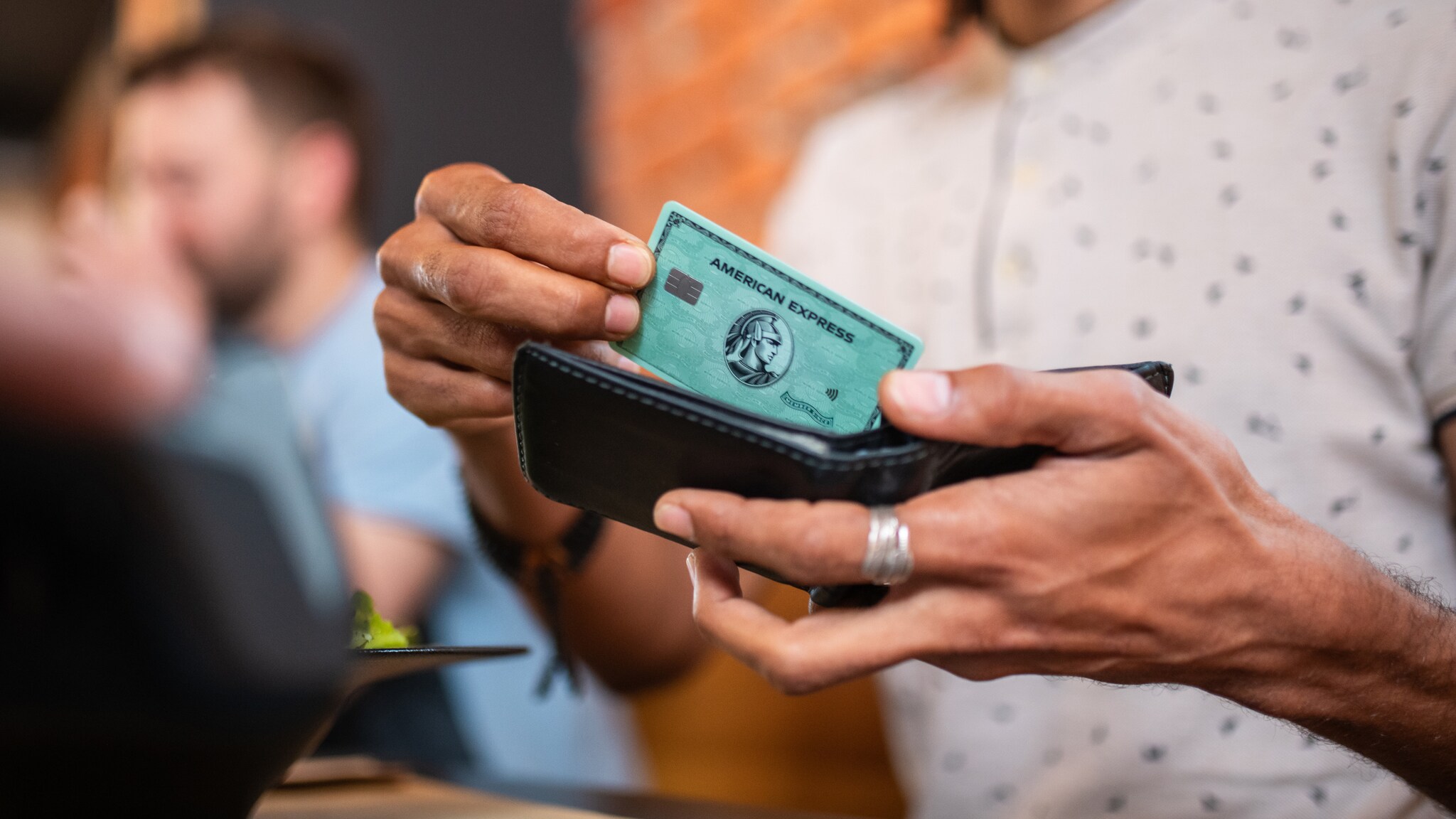 Eine Person zieht eine grüne American Express Kreditkarte aus einer Geldbörse in einem Café.