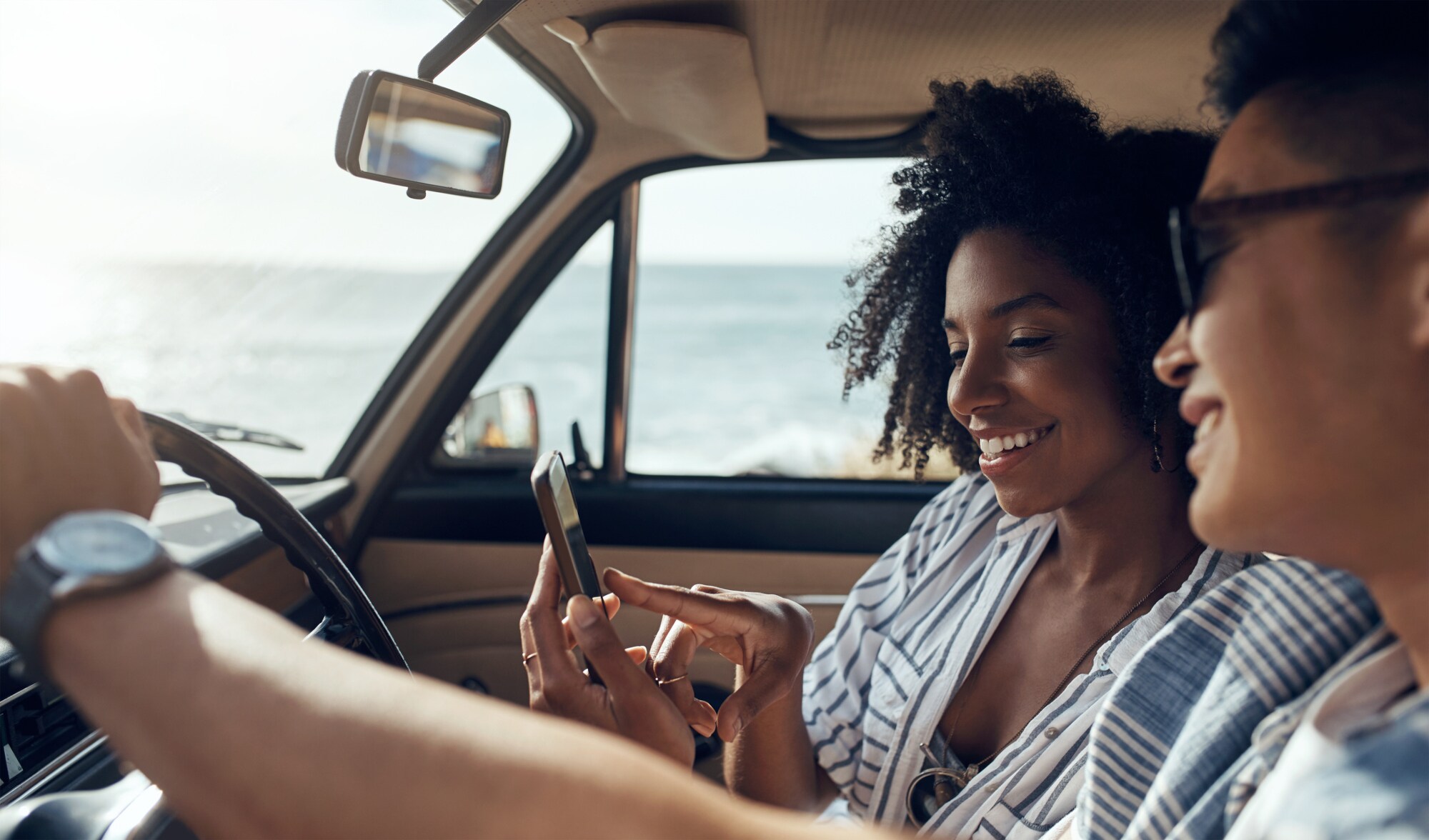 Zwei Frauen auf den Vordersitzen eines Autos schauen während eines spontanen Roadtrips lächelnd auf ein Smartphone, im Hintergrund Meeresküste
