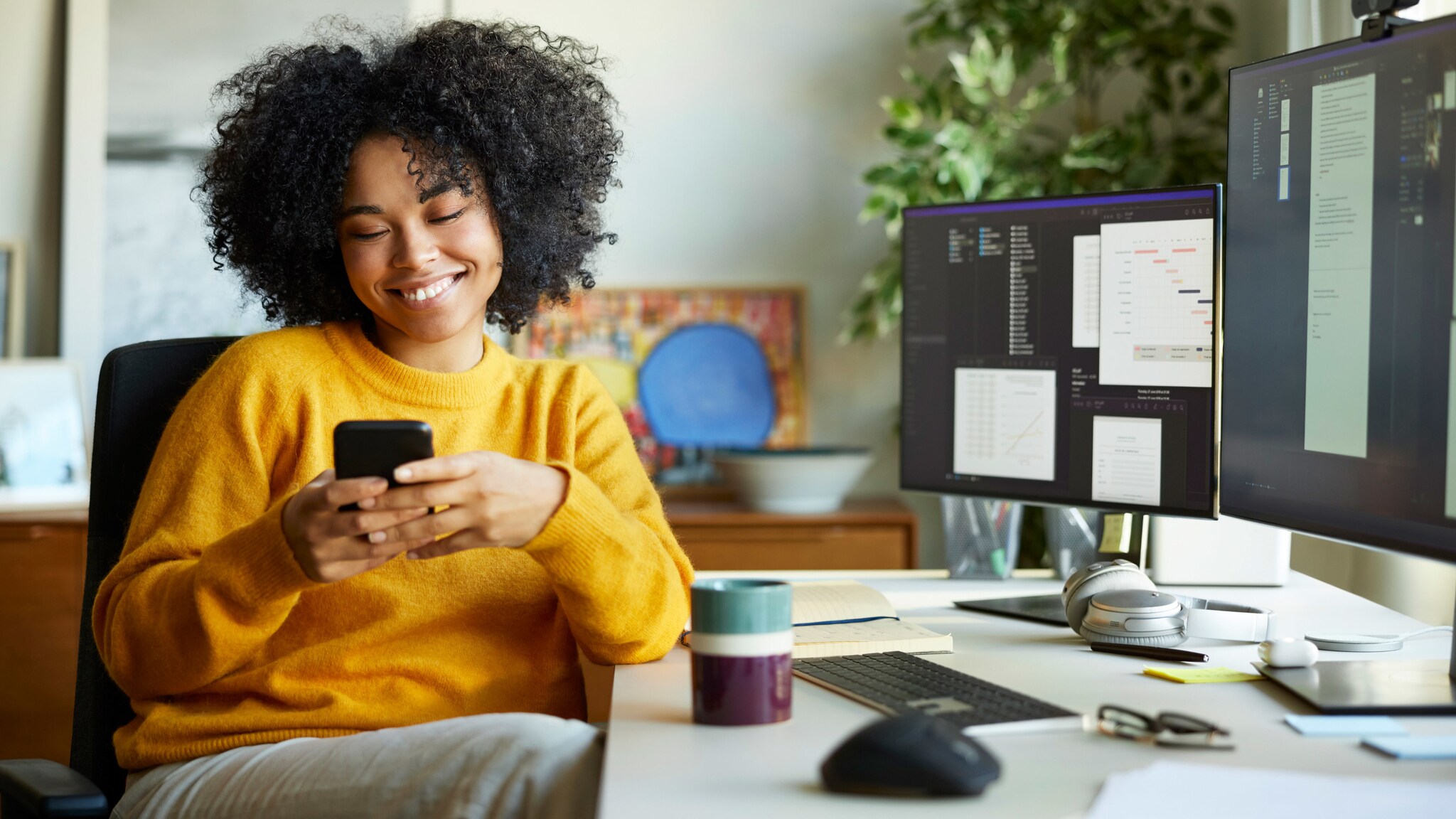 Eine Frau im gelben Pullover sitzt an einem Schreibtisch mit Monitoren und blickt lächelnd auf ein Smartphone in ihren Händen.