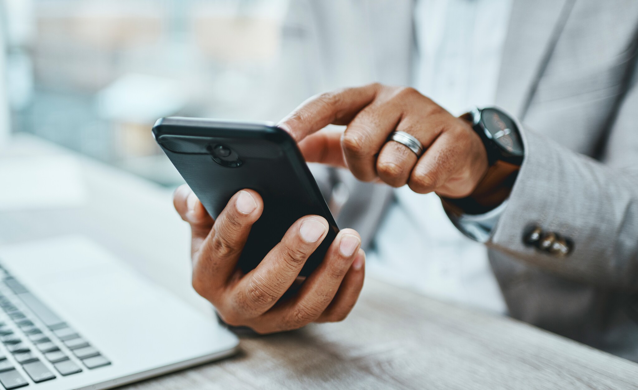 Eine Person hält mir einer Hand ein Smartphone und tippt mit dem Finger der anderen Hand auf das Display über einem Tisch mit Laptop. Eine Person hält mir einer Hand ein Smartphone und tippt mit dem Finger der anderen Hand auf das Display über einem Tisch mit Laptop.