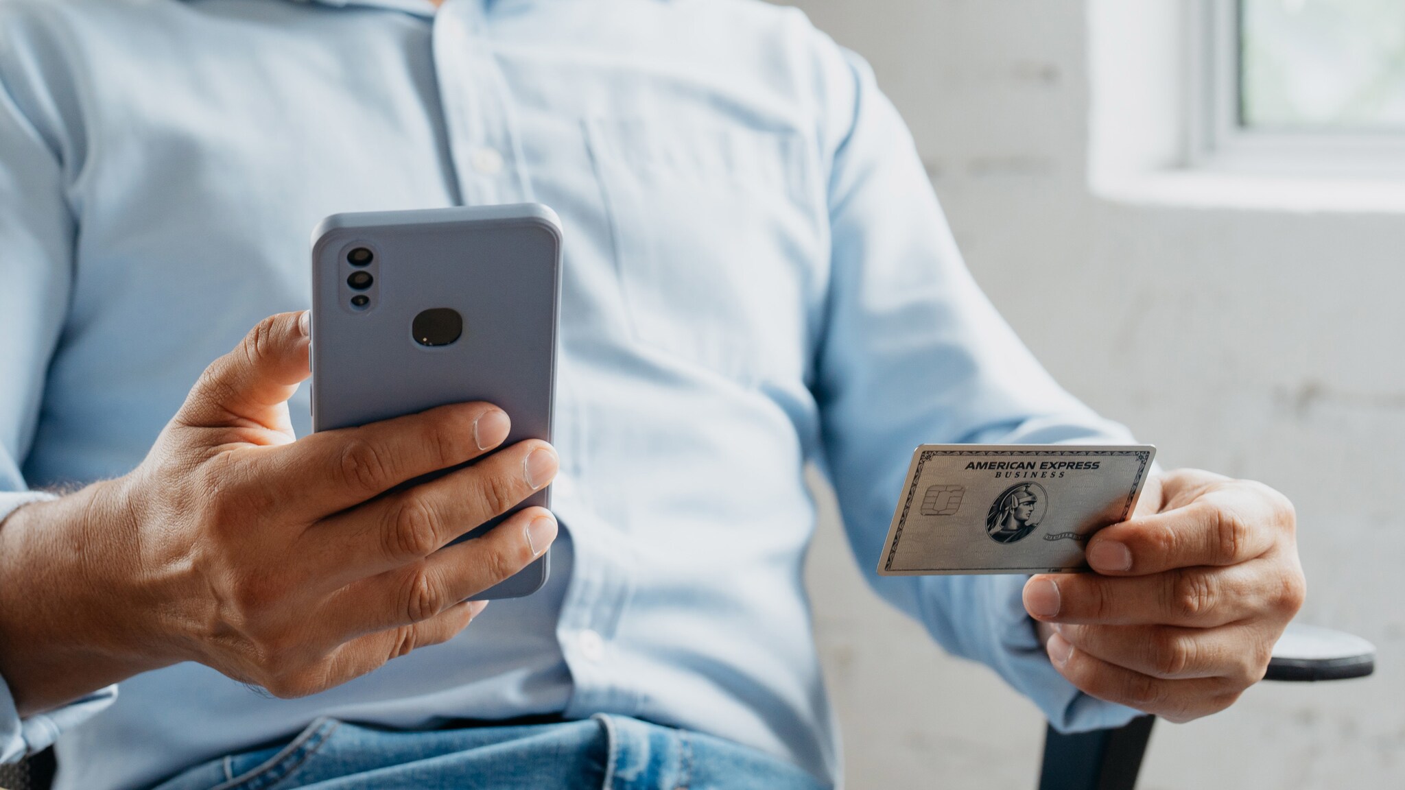 Eine sitzende Person hält in einer Hand ein Smartphone, in der anderen eine Business Platinum Card von American Express. Eine sitzende Person hält in einer Hand ein Smartphone, in der anderen eine Business Platinum Card von American Express.