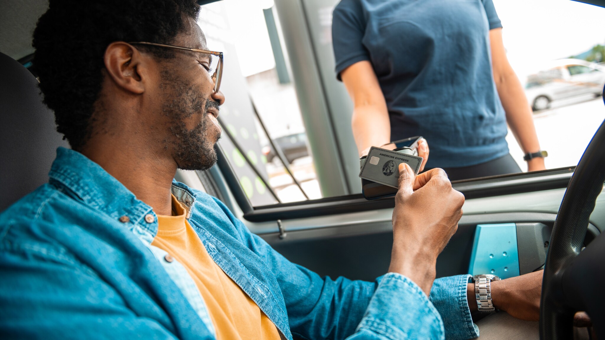 Ein Mann im Jeanshemd am Steuer eines Autos bezahlt kontaktlos mit einer silbernen American Express Kreditkarte an einem Kartenlesegerät, das eine Person in das geöffnete Autofenster hält.