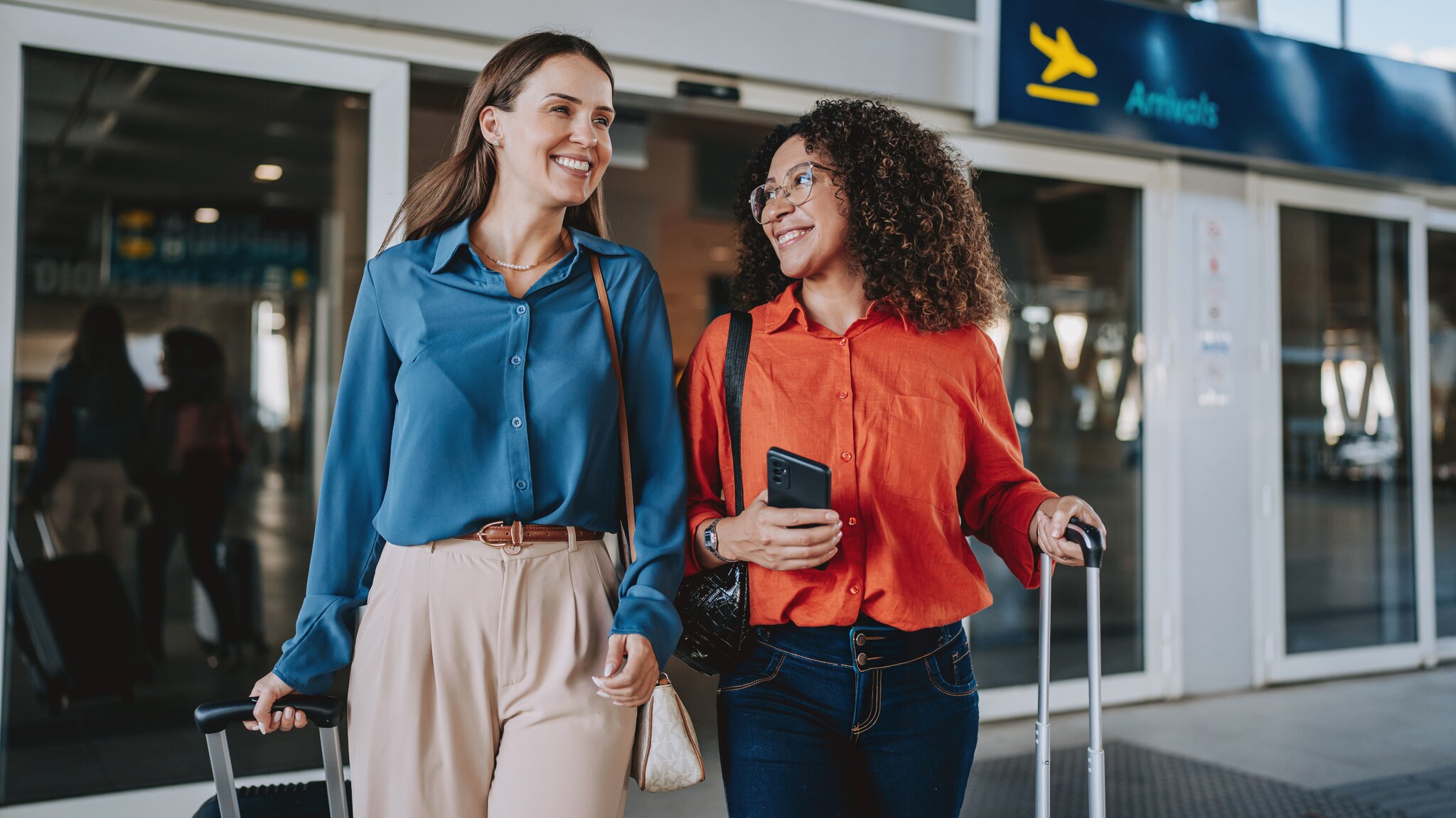 Zwei lachende, elegant gekleidete Frauen mit Trolleys beim Verlassen eines Flughafenterminals. Zwei lachende, elegant gekleidete Frauen mit Trolleys beim Verlassen eines Flughafenterminals.