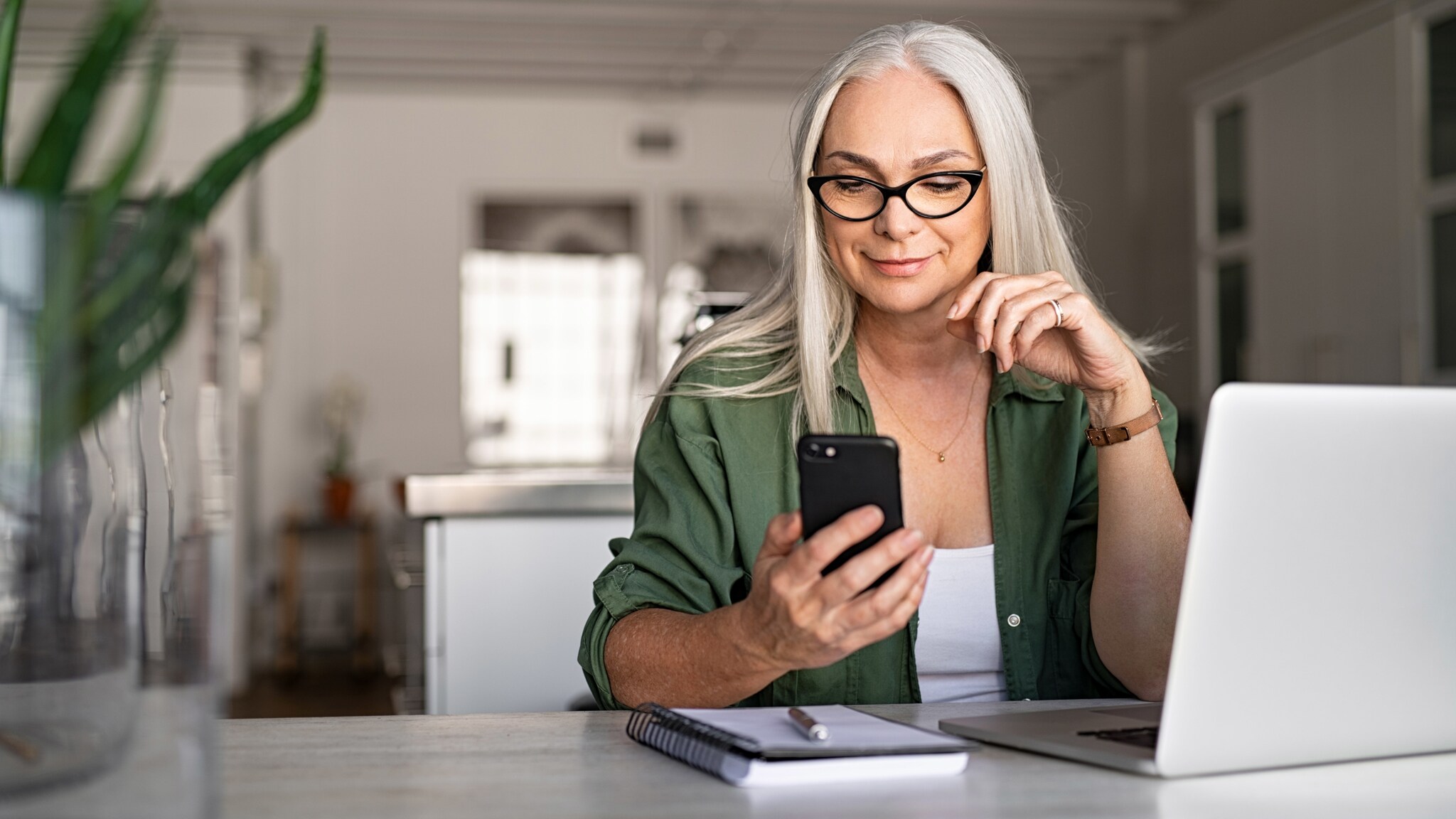 Eine Frau mit langen weißen Haaren sitzt vor einem Laptop an einem Schreibtisch im Büro und schaut zufrieden auf ein Smartphone in ihrer Hand. Eine Frau mit langen weißen Haaren sitzt vor einem Laptop an einem Schreibtisch im Büro und schaut zufrieden auf ein Smartphone in ihrer Hand.
