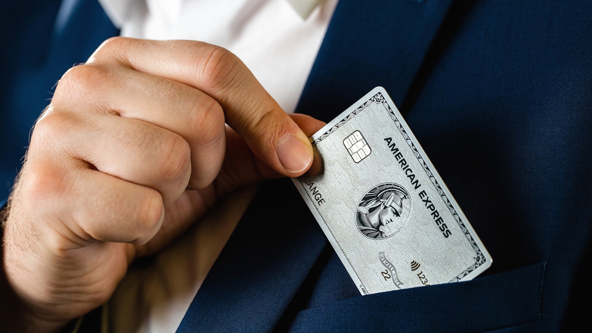 Eine Hand steckt eine silberne Kreditkarte von American Express in die Reverstasche eines blauen Jacketts. Eine Hand steckt eine silberne Kreditkarte von American Express in die Reverstasche eines blauen Jacketts.