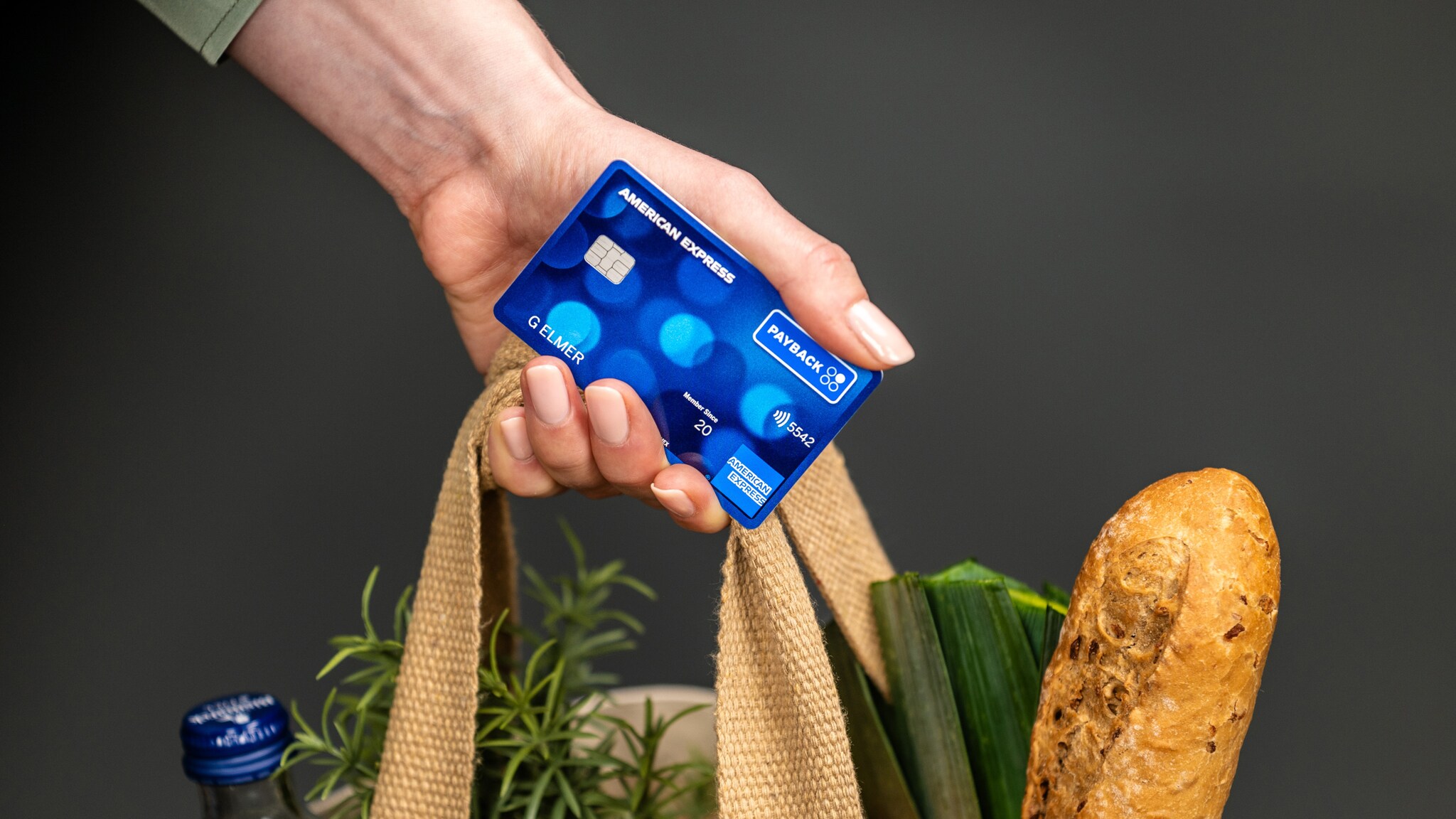 Eine blaue Payback Kreditkarte von American Express in einer Hand, die ebenfalls die Griffe einer Tasche mit Lebensmitteleinkäufen hält.