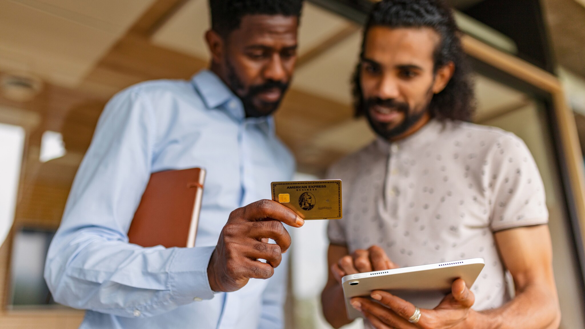 Zwei stehende Männer schauen gemeinsam auf ein Tablet, das einer in den Händen hält, und eine Business Gold Card von American Express, die der andere hält.