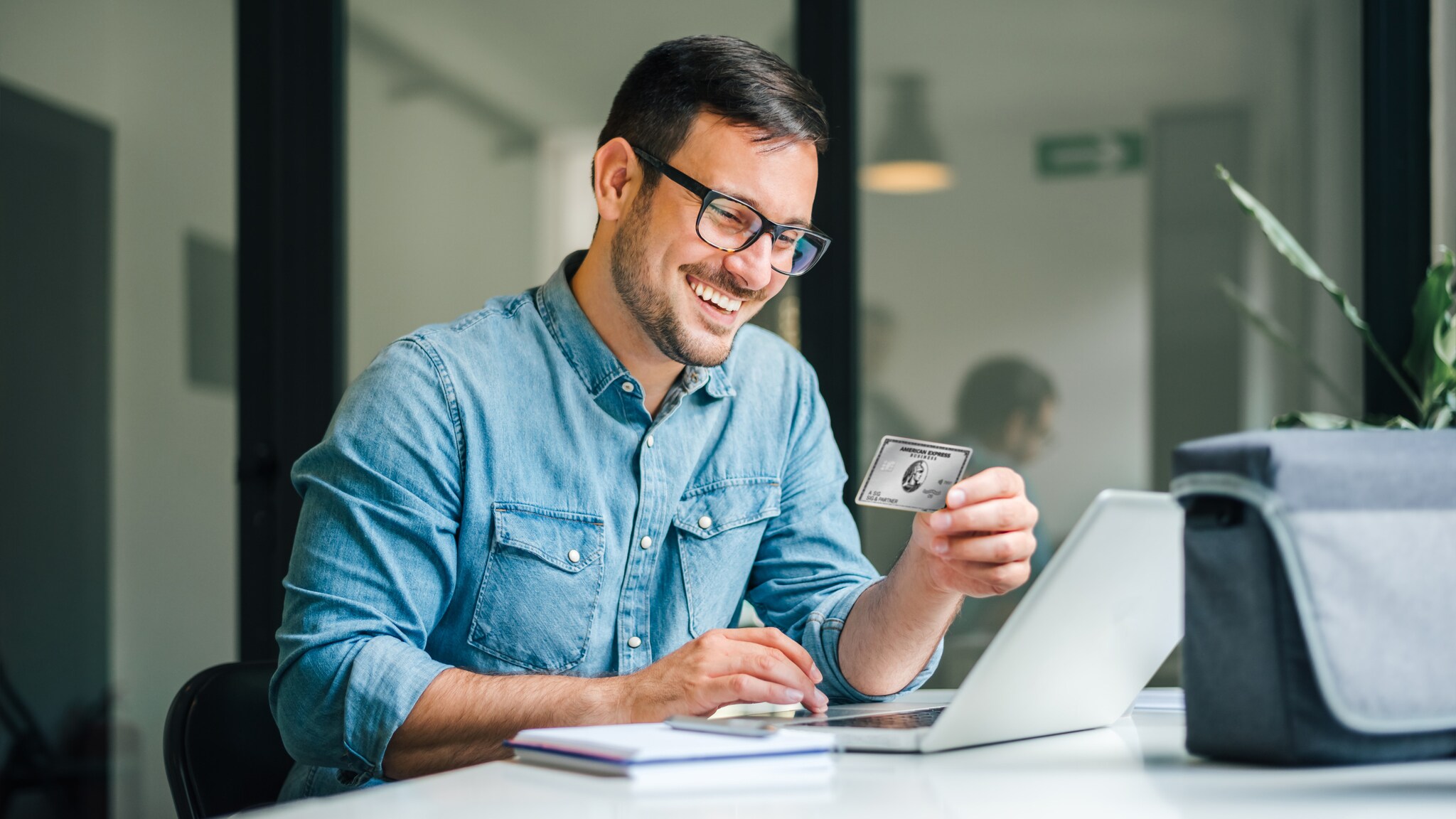 Ein lächelnder Mann im Jeanshemd sitzt vor einem Laptop und hält eine Business Platinum Card von American Express in der Hand.