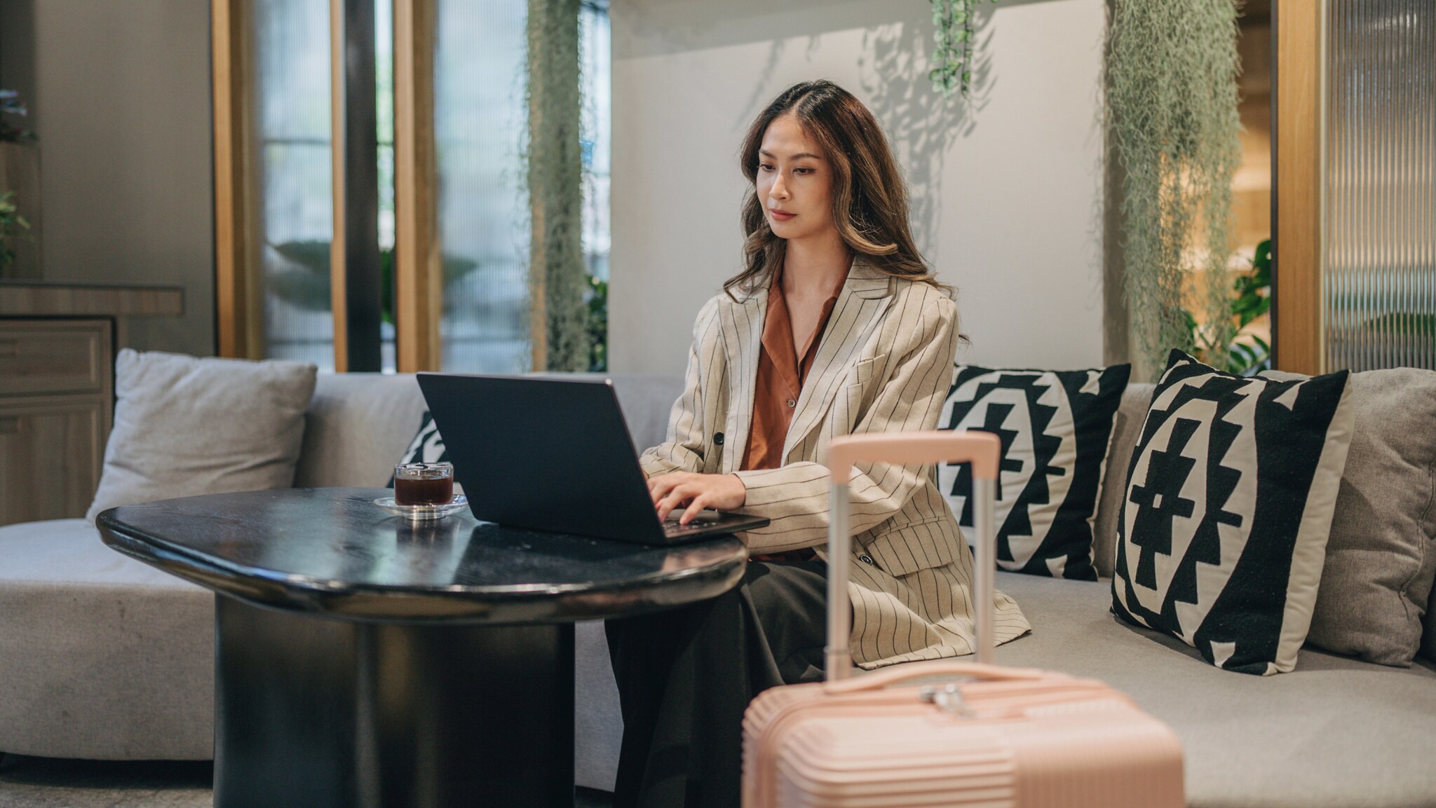 Eine junge Frau sitzt auf einem Sofa an einem Tisch und einem Laptop, neben ihr ein Hartschalenkoffer.