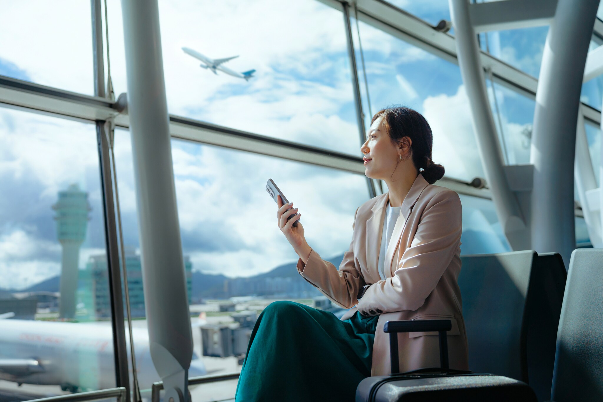 Eine moderne Geschäftsfrau sitzt mit einem Smartphone in ihrer Hand an einem Gate am Flughafen und blickt aufs Rollfeld neben ihr steht ein Trolley. Eine moderne Geschäftsfrau sitzt mit einem Smartphone in ihrer Hand an einem Gate am Flughafen und blickt aufs Rollfeld neben ihr steht ein Trolley.