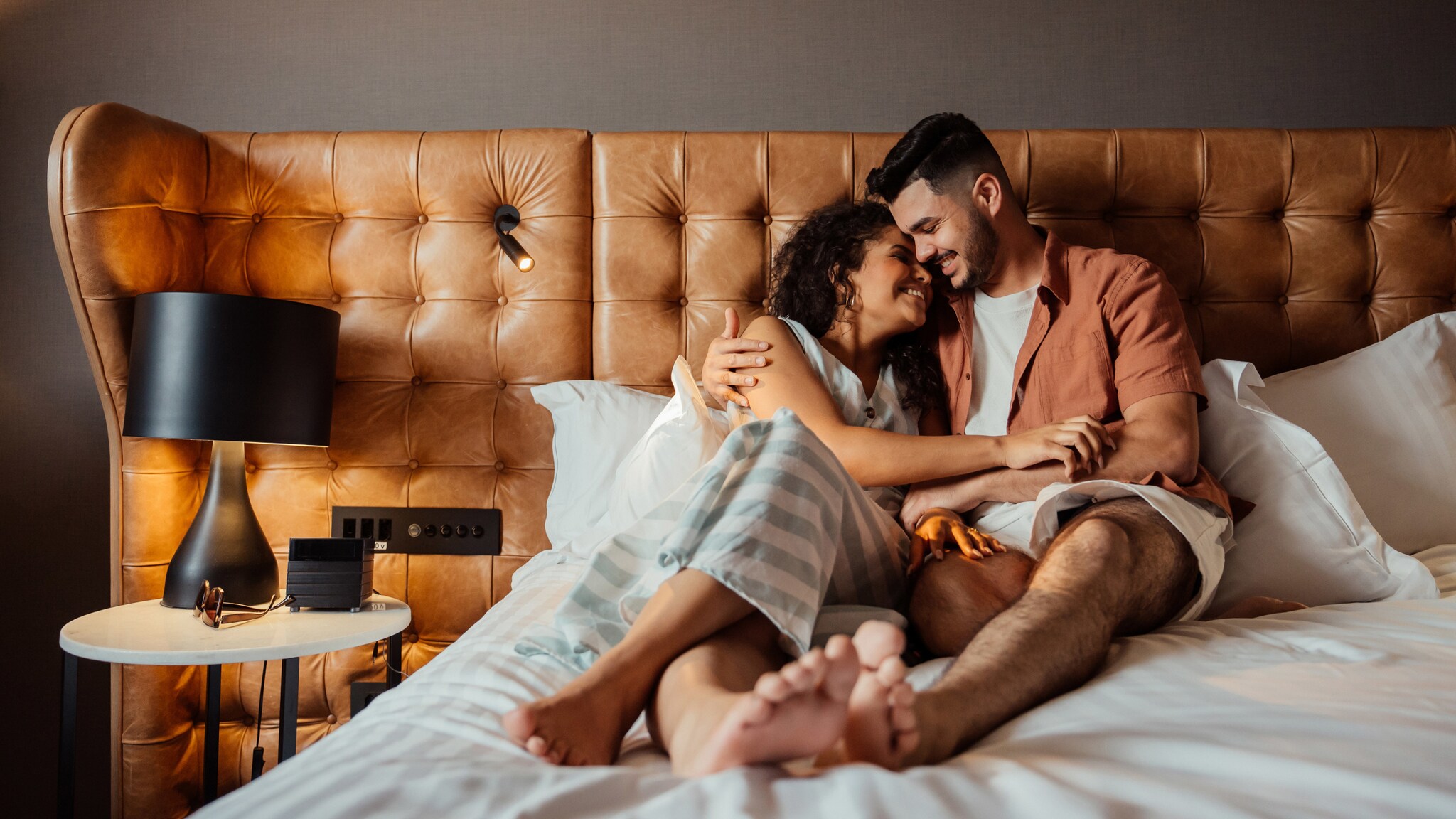 Ein glückliches Paar liegt kuschelnd in einem Hotelbett bei Tageslicht.