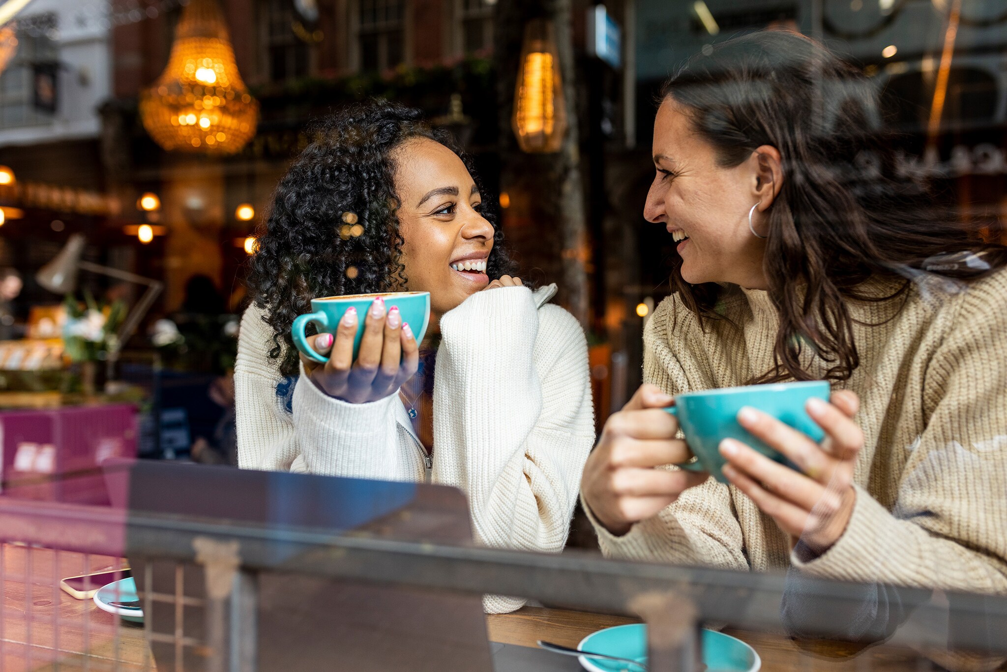 Zwei Frauen mit Kaffeetassen in den Händen sitzen am Fenster eines Cafés und schauen sich fröhlich an.