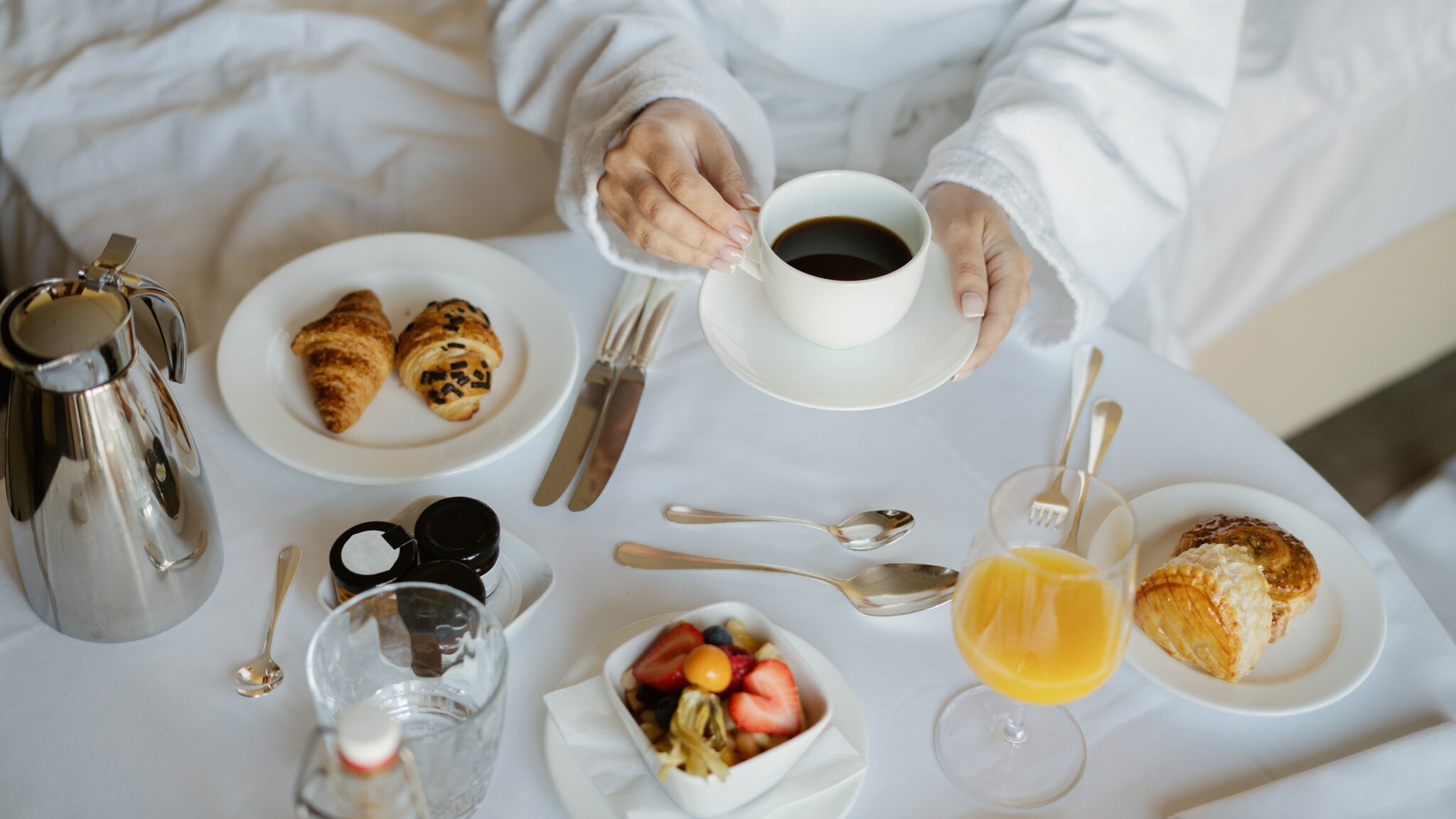Zwei Hände einer Person in einem weißen Bademantel umgreifen eine Kaffeetasse auf einem vornehm eingedeckten Frühstückstisch an einem Bett.