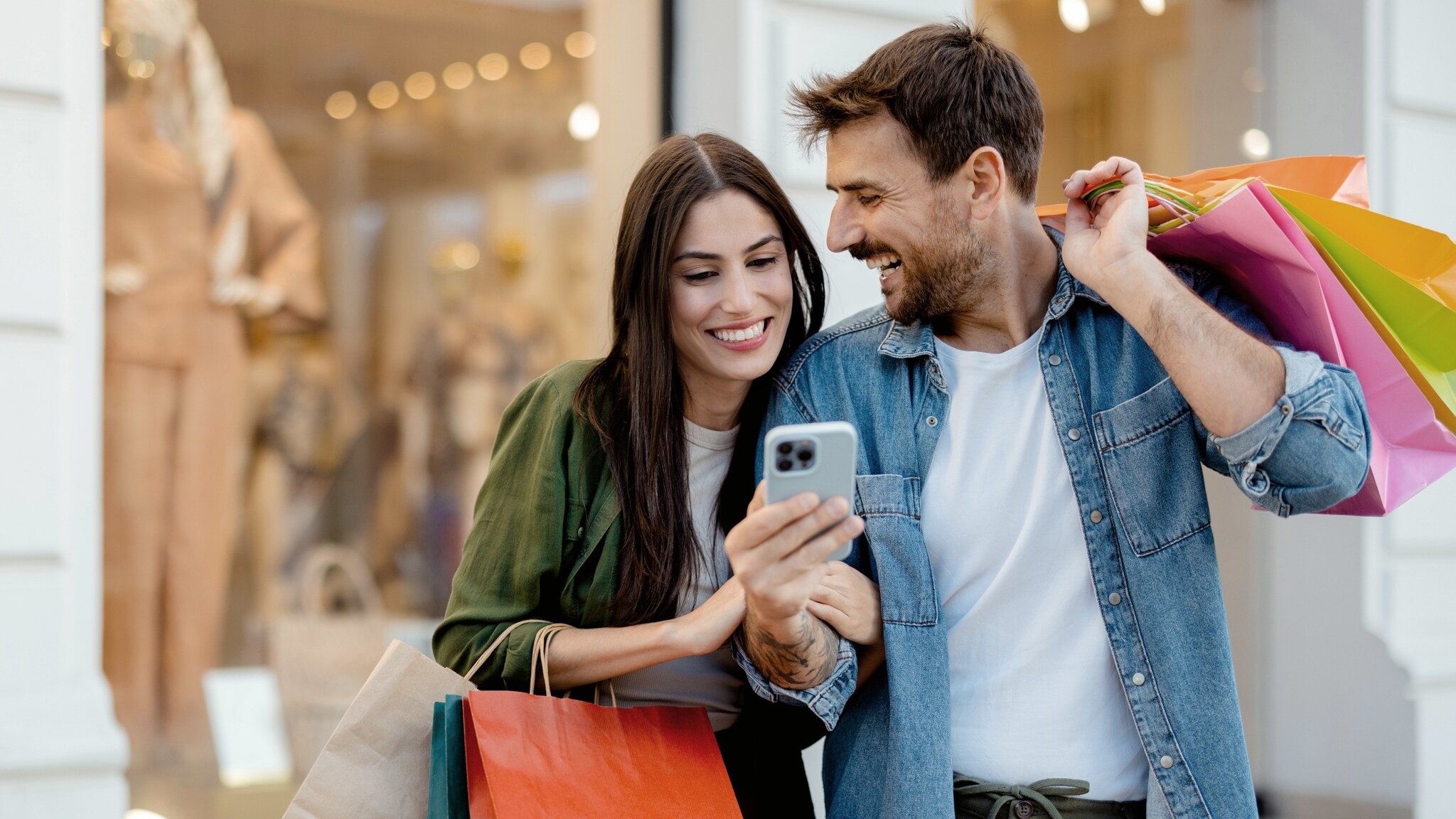 Eine Frau und ein Mann stehen eingehakt und gut gelaunt vor einem Modegeschäft mit bunten Einkaufstüten in den Händen, vor ihnen ein hochgehaltenes Smartphone in seiner Hand. Eine Frau und ein Mann stehen eingehakt und gut gelaunt vor einem Modegeschäft mit bunten Einkaufstüten in den Händen, vor ihnen ein hochgehaltenes Smartphone in seiner Hand.