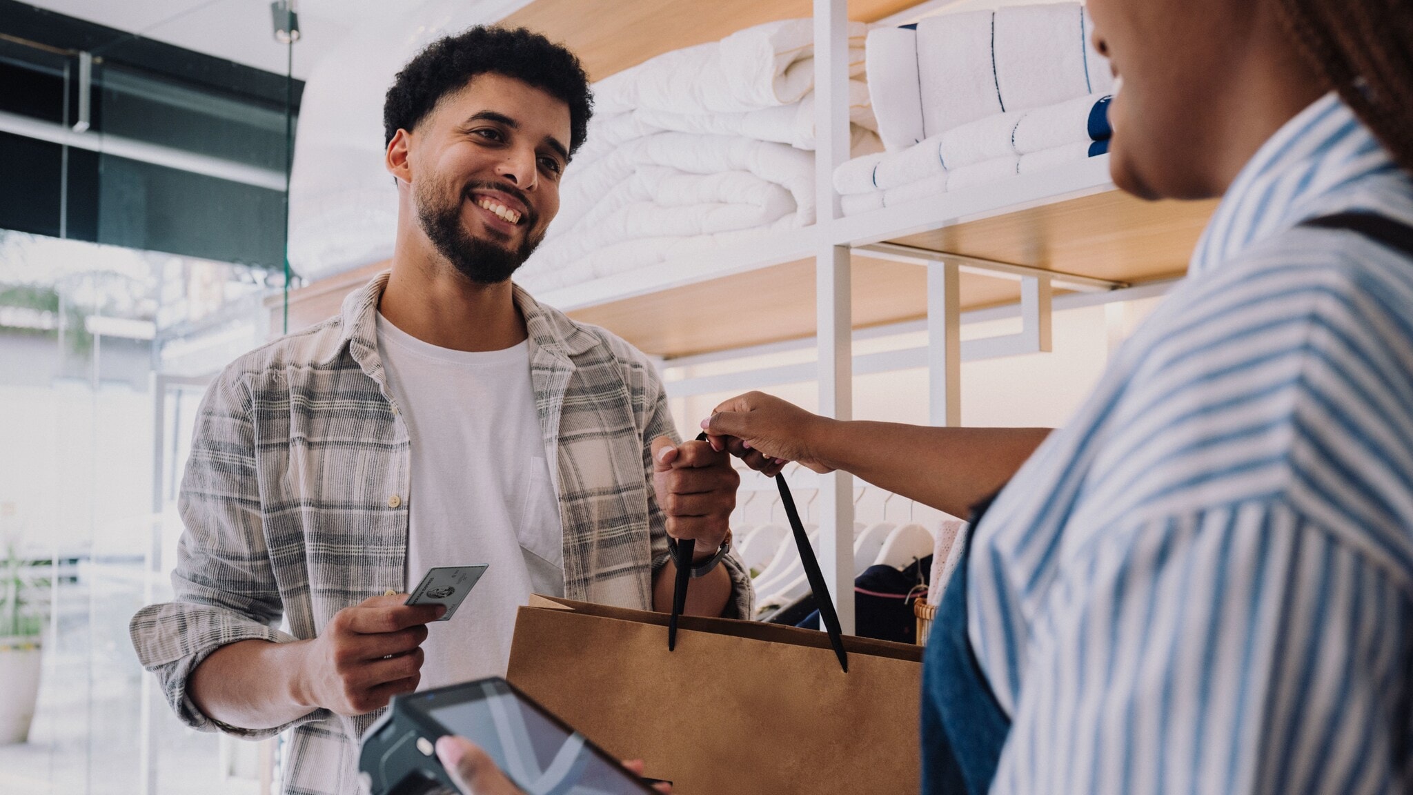 Ein Mann nimmt lächelnd eine Einkaufstüte von einer Verkäuferin in einem Geschäft entgegen, dabei hält er eine silberne Kreditkarte von American Express in der Hand. Ein Mann nimmt lächelnd eine Einkaufstüte von einer Verkäuferin in einem Geschäft entgegen, dabei hält er eine silberne Kreditkarte von American Express in der Hand.