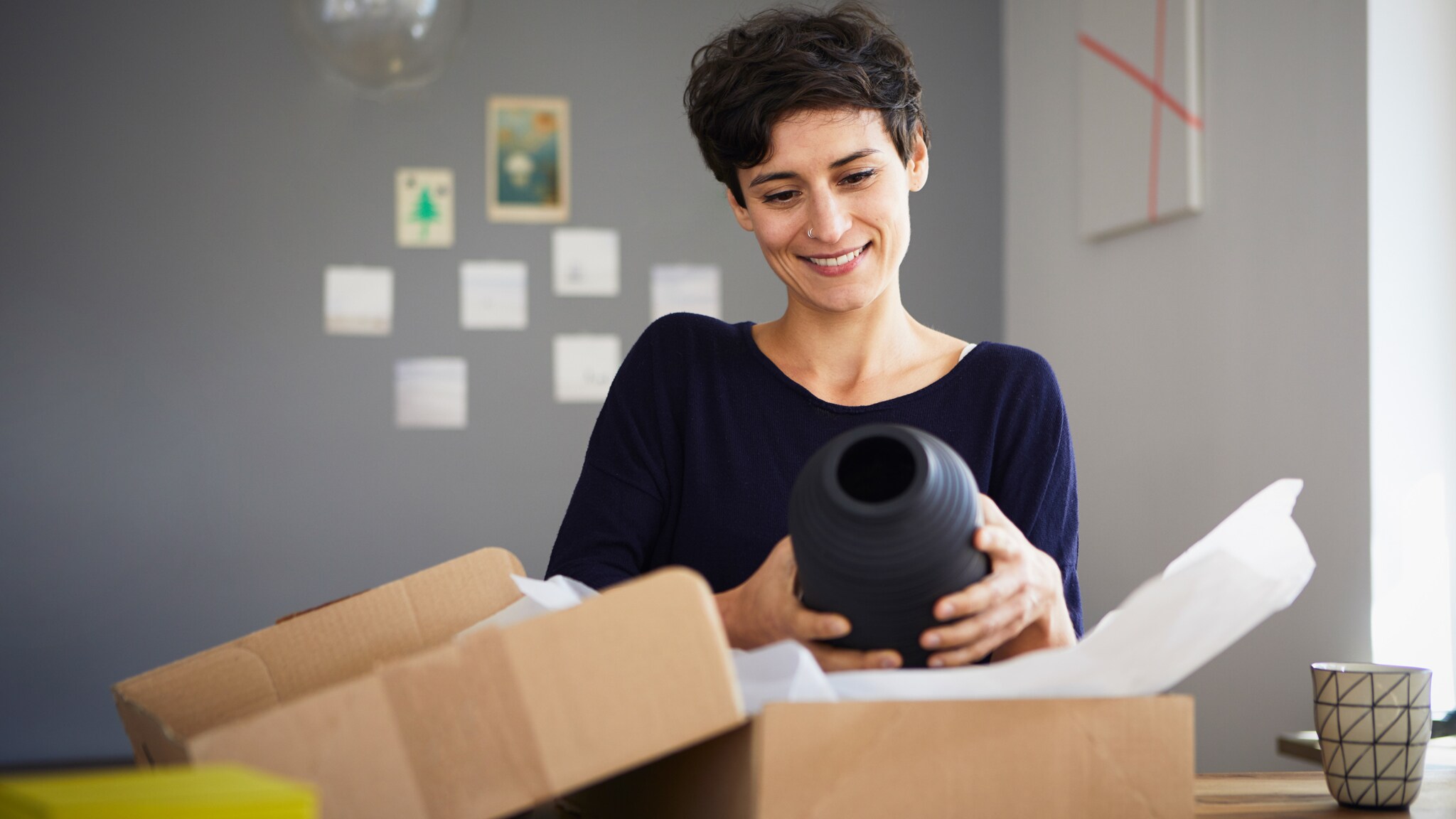 Eine lächelnde Frau hält eine schwarze Vase über ein Paket mit Packpapier auf einem Tisch.