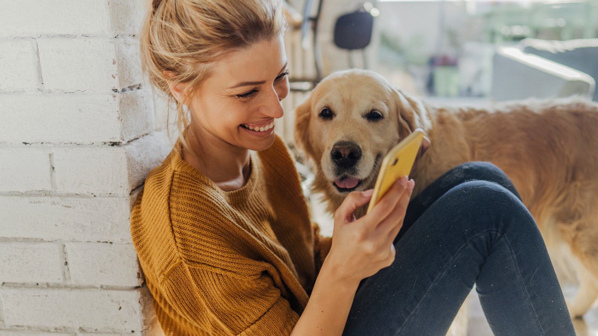 Eine junge Frau sitzt in einer Wohnung neben einem Golden Retriever auf dem Boden und schaut gut gelaunt auf ihr Smartphone.