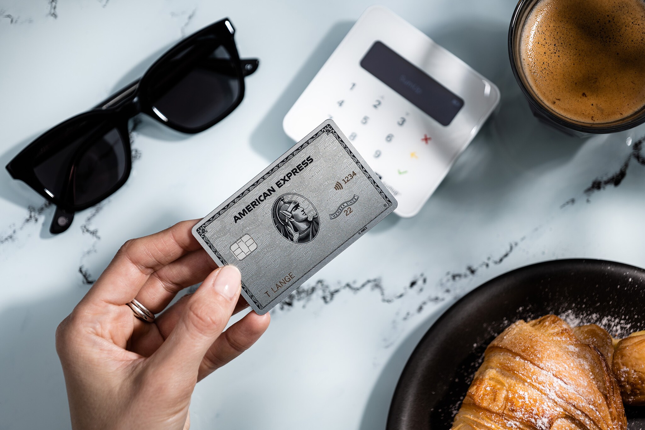 Kontaktlose Bezahlung mit einer Amex Platinum Card an einem Kartenlesegerät auf einem Tisch neben Kaffee, Gebäck und Sonnenbrille. Kontaktlose Bezahlung mit einer Amex Platinum Card an einem Kartenlesegerät auf einem Tisch neben Kaffee, Gebäck und Sonnenbrille.
