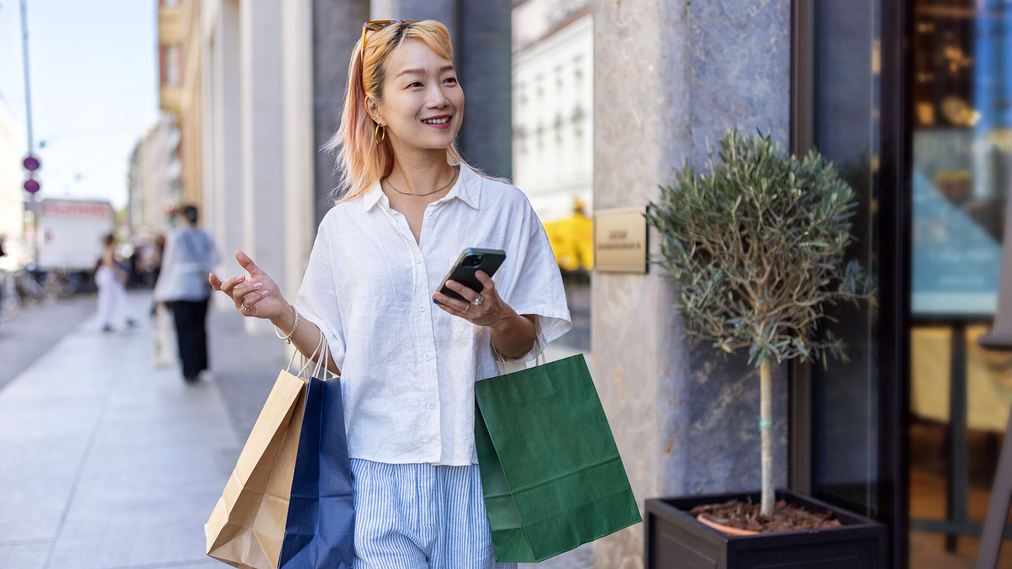 Eine junge Frau mit Hut steht in einer Innenstadt und hält ein Smartphone und mehrere Einkaufstüten in den Händen. Eine junge asiatische Frau mit blonden Haaren mit Einkaufstüten und Smartphone in den Händen geht lächelnd über eine Einkaufsstraße.