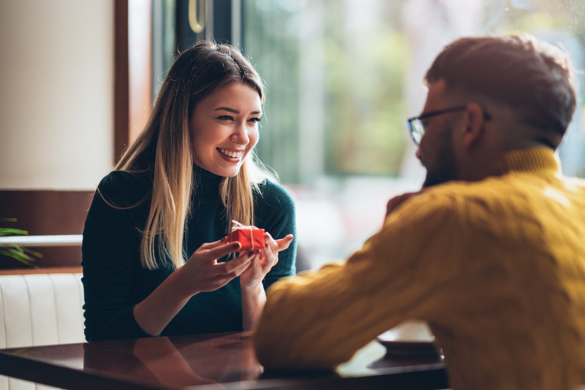 Eine junge Frau sitzt einem Mann in einem Café gegenüber und hält lächelnd eine kleine, rote Geschenkbox in ihren Händen