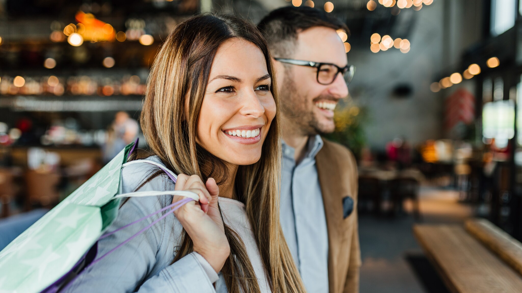 Eine Frau hält eine Tasche über der Schulter, neben ihr steht ein Mann, beide lächeln.