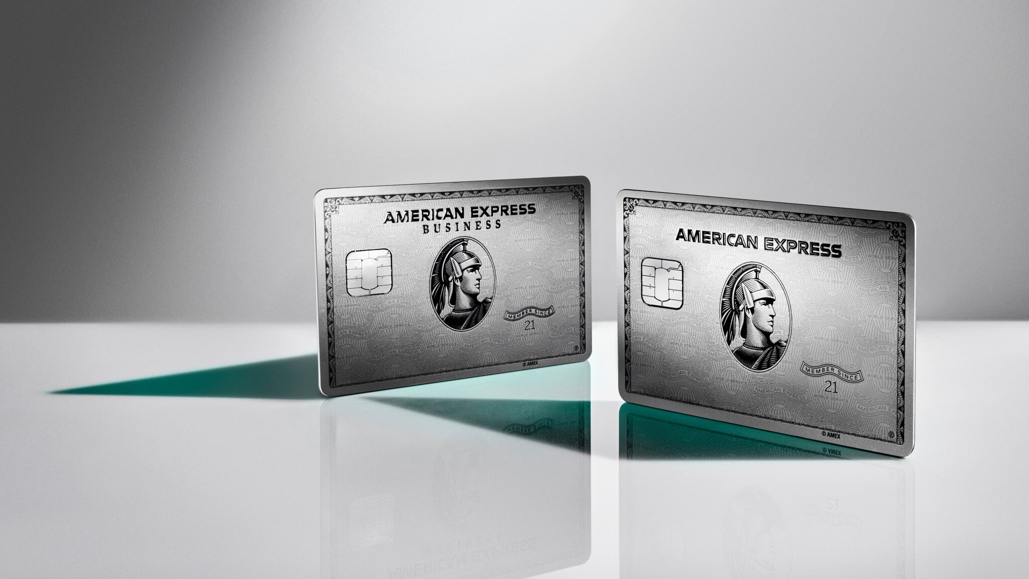 Die American Express Business Platinum Card neben der Platinum Plus Card, aufgestellt auf einer glänzenden Fläche vor grauem Hintergrund.