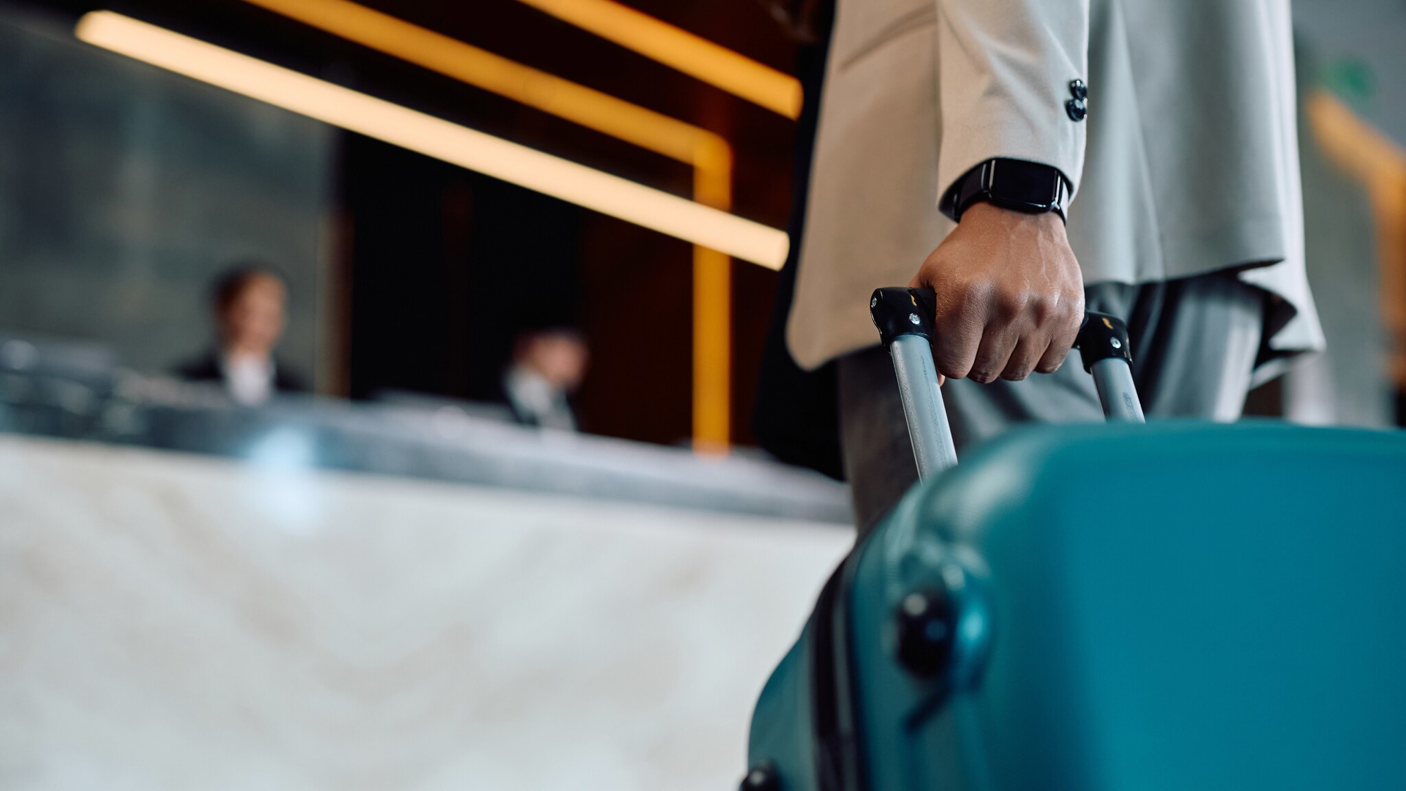 Nahaufnahme einer Hand am Griff eines türkisfarbenen Hartschalenkoffers, dahinter eine Hotelrezeption in Unschärfe.