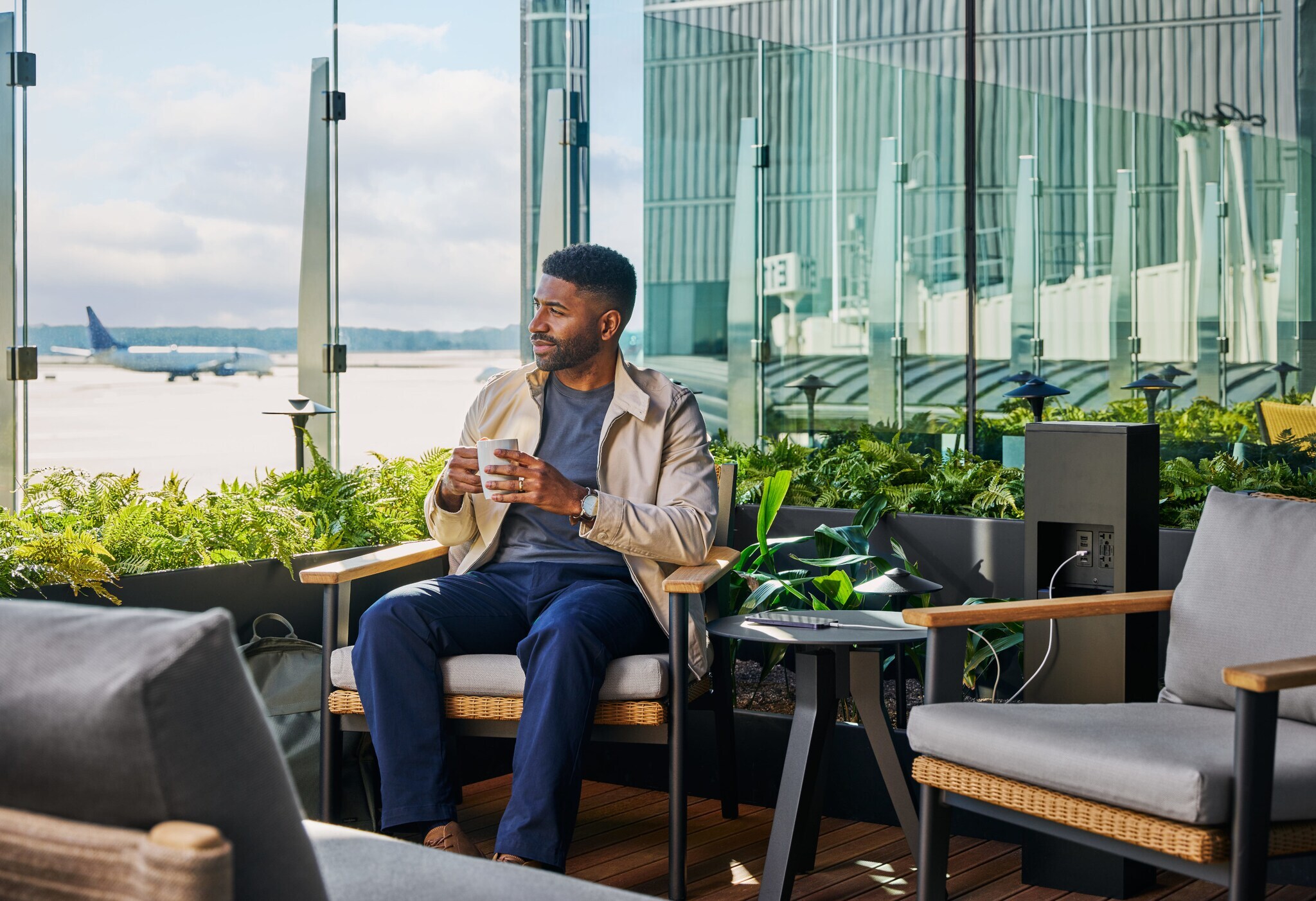 Ein Mann sitzt mit einer Kaffeetasse auf einer Terrasse mit Blick auf ein Flughafenrollfeld.