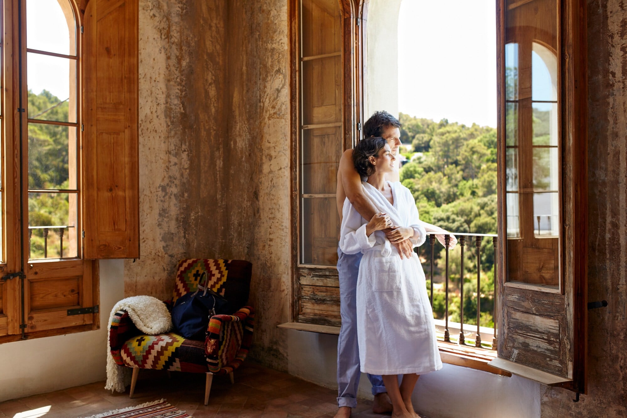 Ein sich umarmendes Paar steht an einem großen Fenster in einer mediterranen Hotelsuite und schaut in die grüne Landschaft; sie trägt einen weißen Bademantel Ein sich umarmendes Paar steht an einem großen Fenster in einer mediterranen Hotelsuite und schaut in die grüne Landschaft; sie trägt einen weißen Bademantel
