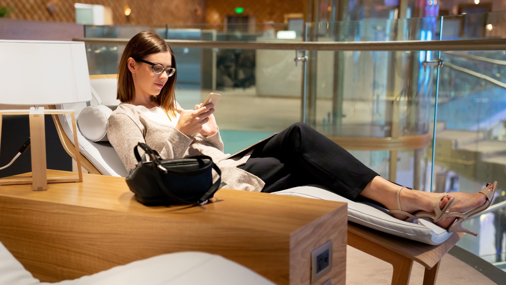 Eine elgant gekleidete Frau liegt auf einer Liege in einer Flughafenlounge und schaut auf ihr Smartphone. Eine elgant gekleidete Frau liegt auf einer Liege in einer Flughafenlounge und schaut auf ihr Smartphone.