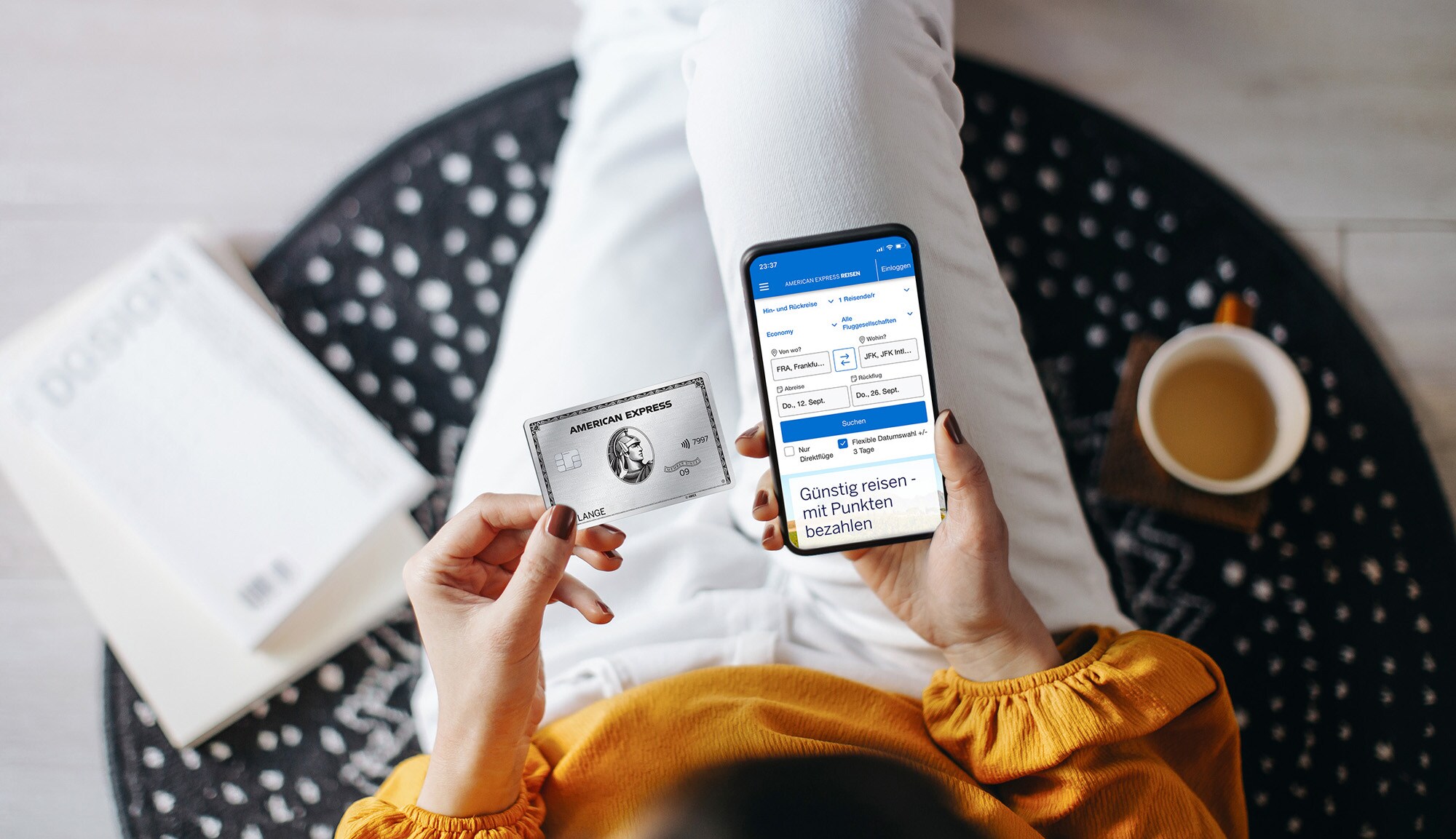 Aufsicht einer sitzenden Person mit silberner Kreditkarte von American Express und Smartphone mit Amex Flugbuchungsmaske auf dem Display in ihren Händen. Aufsicht einer sitzenden Person mit silberner Kreditkarte von American Express und Smartphone mit Amex Flugbuchungsmaske auf dem Display in ihren Händen.
