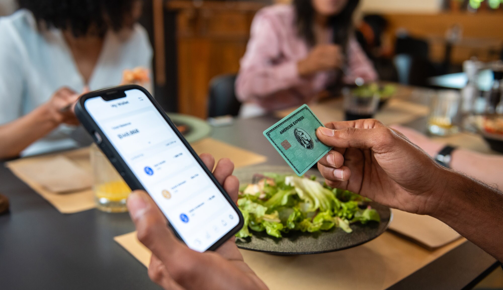 Eine Person hält eine grüne American Express Kreditkarte und ein Smartphone mit Amex App in den Händen an einem Tisch beim Mittagessen.