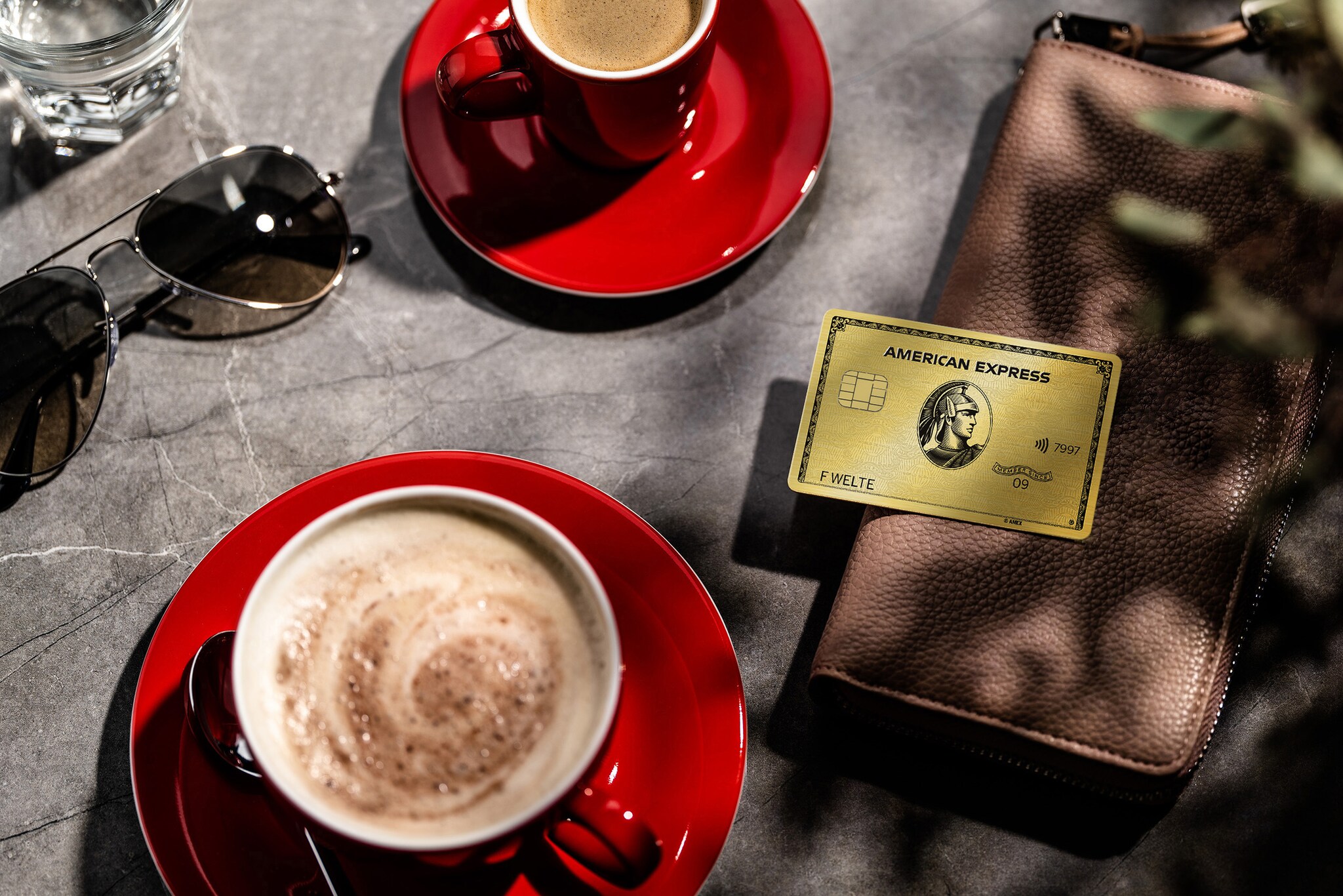 Eine goldene American Express Kreditkarte liegt auf einem Portemonnaie neben zwei roten Kaffeetassen auf einem Tisch.