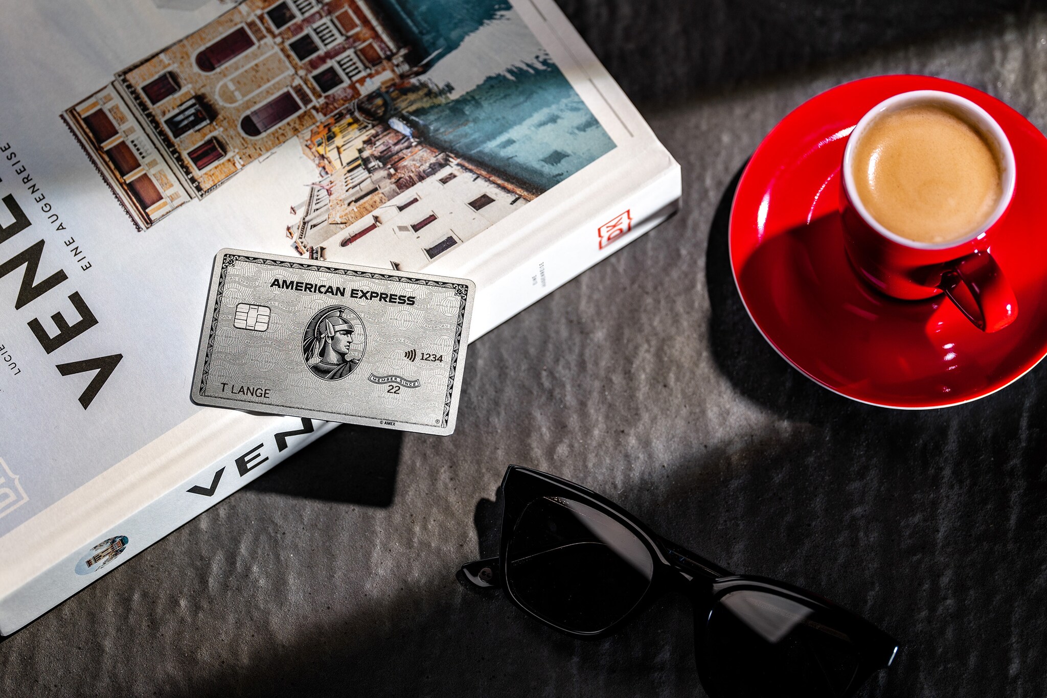 Eine silberne Kreditkarte von American Express liegt auf einem Reisebuch neben einer Espressotasse und einer Sonnenbrille. Eine silberne Kreditkarte von American Express liegt auf einem Reisebuch neben einer Espressotasse und einer Sonnenbrille.