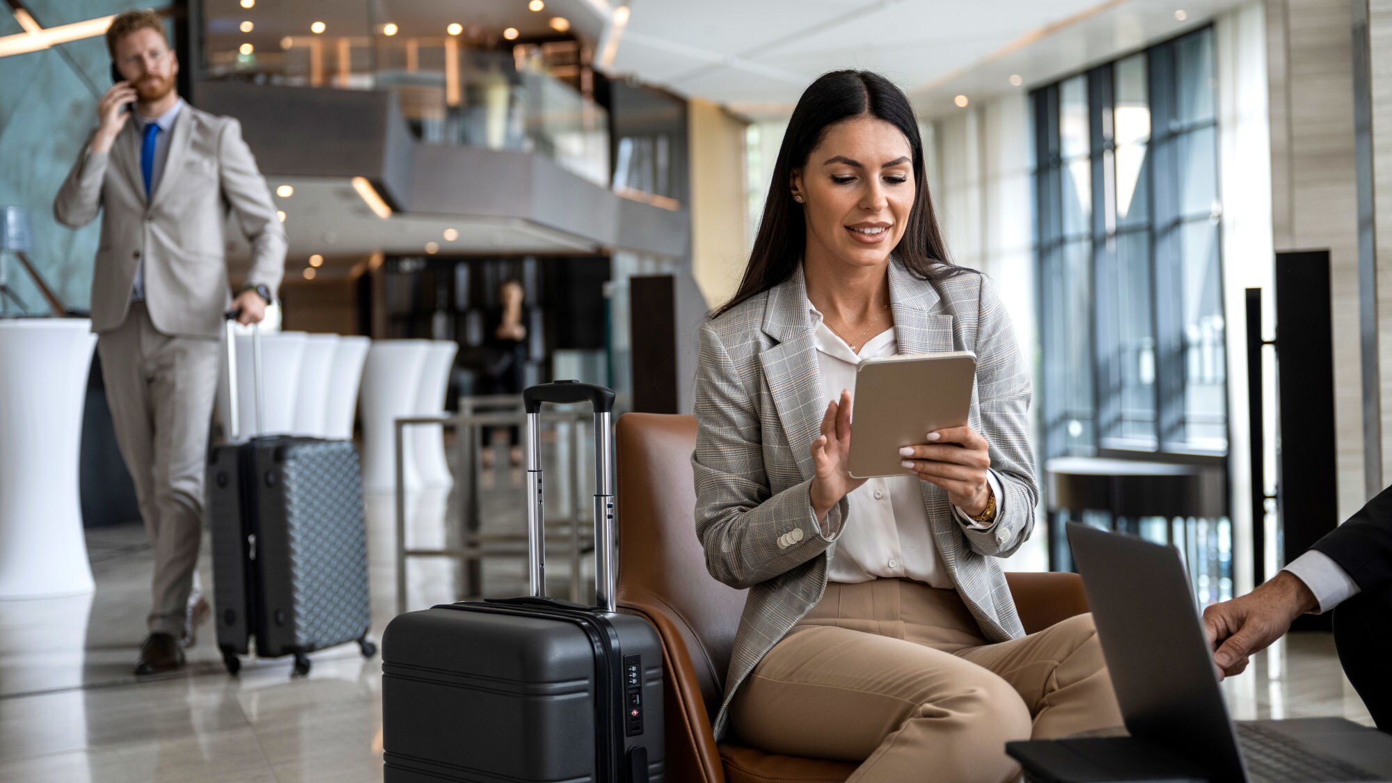 Eine Frau in heller Kleidung sitzt in einer Flughafen-Lounge und nutzt ein Tablet, im Hintergrund geht ein Mann mit Telefon am Ohr im grauen Anzug mit Trolley.