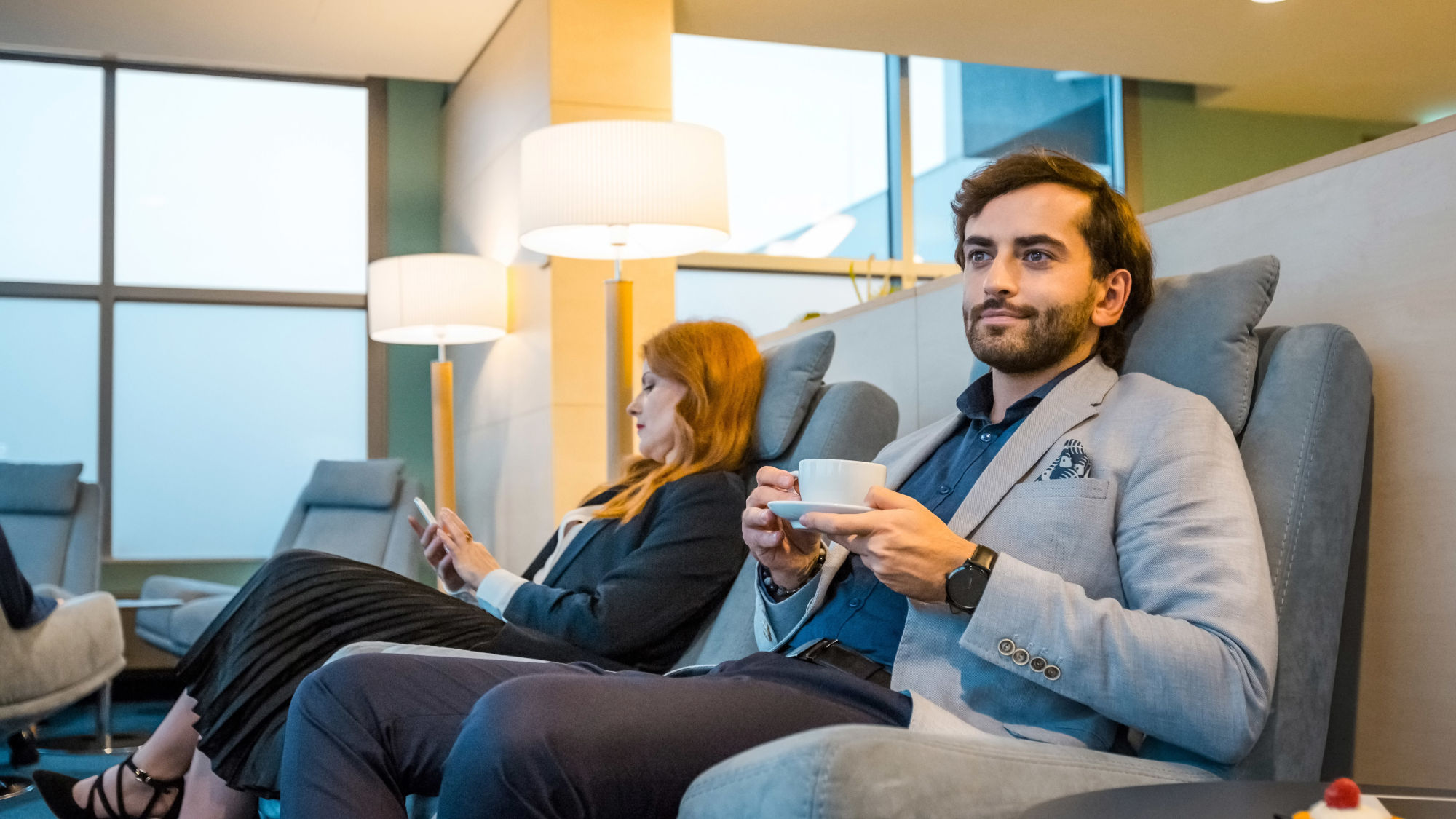 Zwei elegant gekleidete Personen sitzen entspannt in Sesseln einer Flughafen-Lounge mit Panoramafenster, eine hält eine Tasse Kaffee, die andere schaut auf ihr Smartphone.
