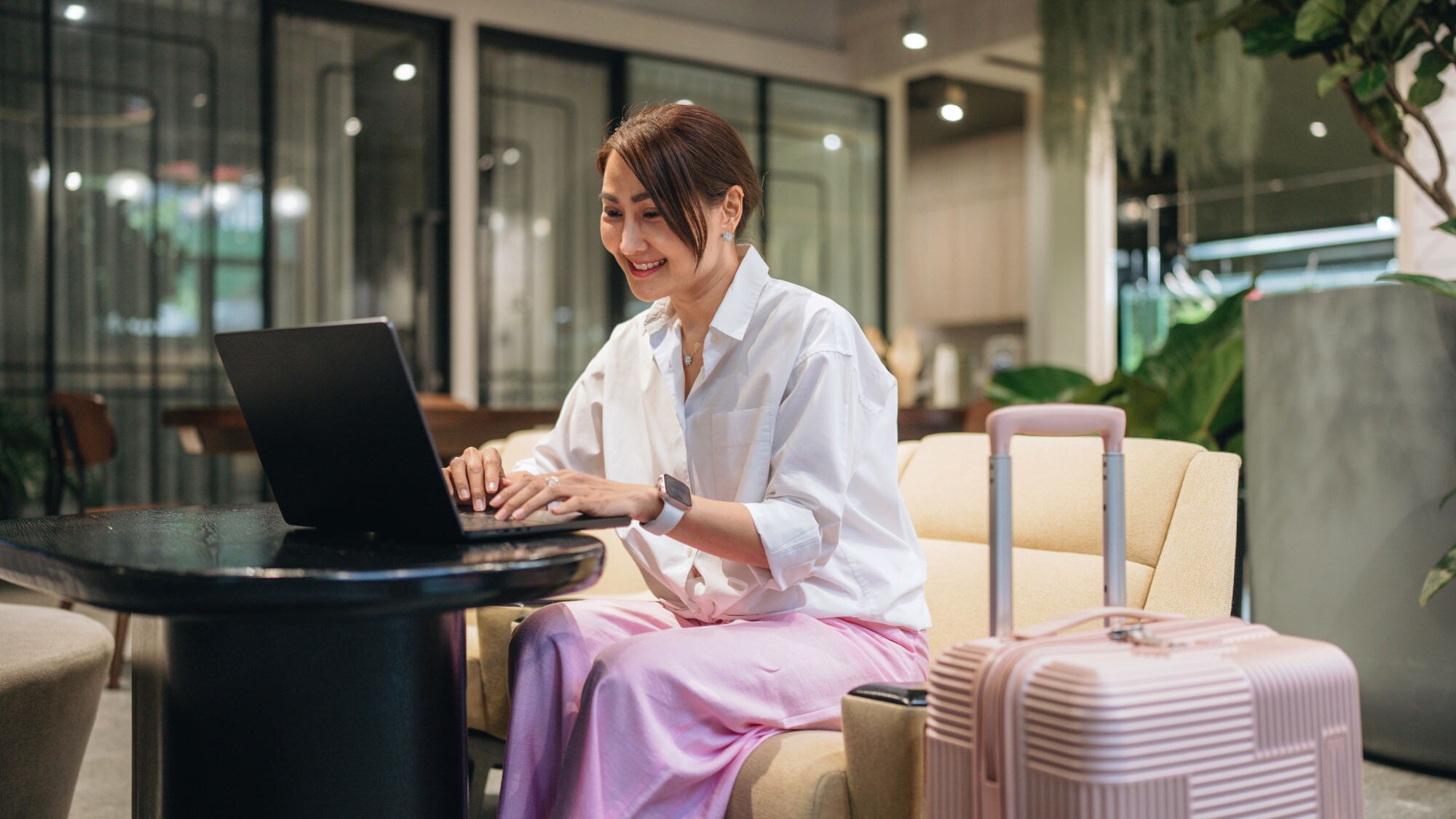 Eine Frau sitzt lächelnd an einem Laptop in einer Lounge, neben ihr ein großer rosafarbener Hartschalenkoffer. Eine Frau sitzt lächelnd an einem Laptop in einer Lounge, neben ihr ein großer rosafarbener Hartschalenkoffer.