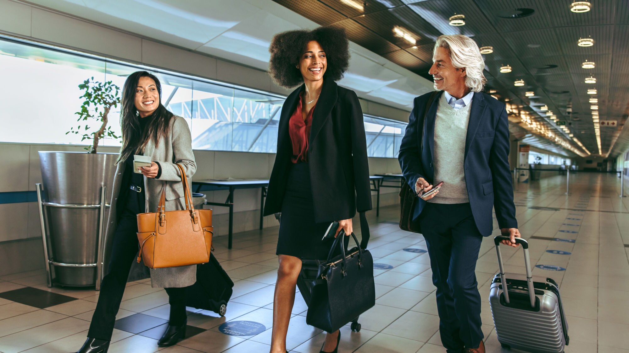 Drei Personen in eleganter Business-Kleidung und Handgepäck laufen nebeneinander durch ein Flughafenterminal. Drei Personen in eleganter Business-Kleidung und Handgepäck laufen nebeneinander durch ein Flughafenterminal.